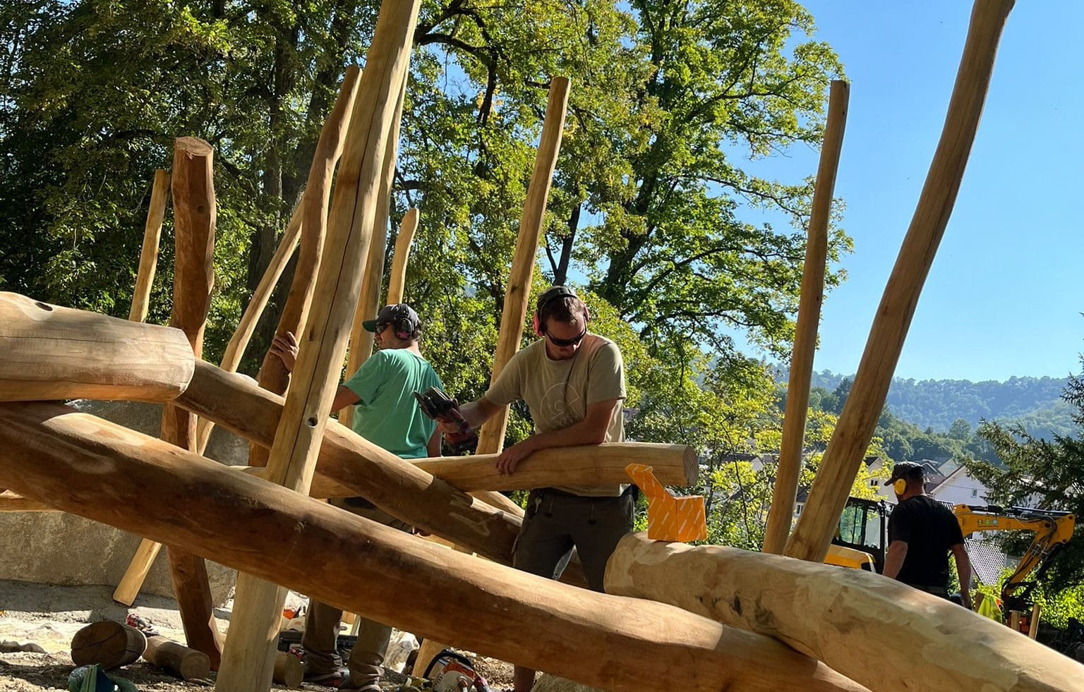 Zwei Personen errichten im Freien, umgeben von Bäumen, eine Holzkonstruktion. Sie arbeiten unter klarem Himmel mit Baumstämmen und Balken.