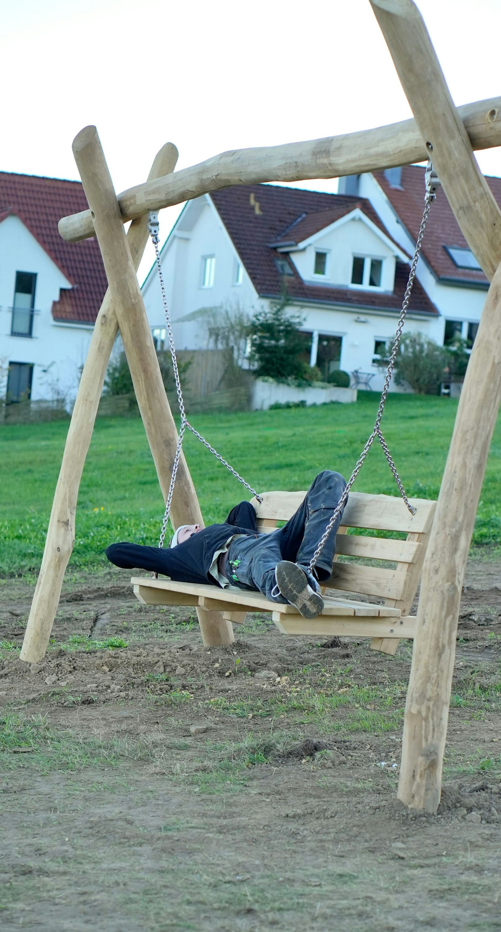Person, die auf einer Holzschaukel in einer Grasfläche mit Häusern im Hintergrund liegt.
