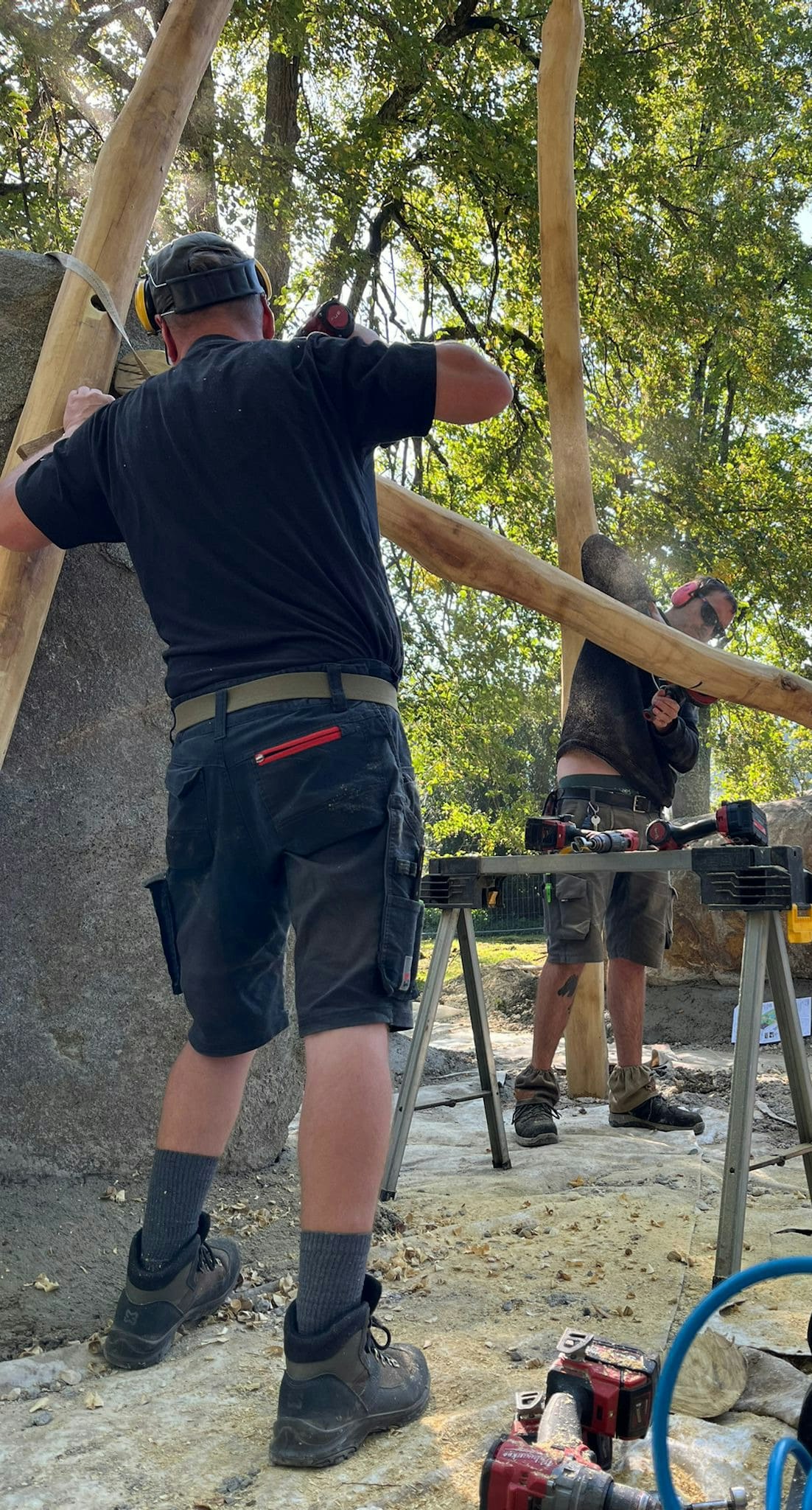 Zwei Männer arbeiten im Freien mit Werkzeugen und Sägen an der Errichtung einer Holzkonstruktion. Die Szene ist von Bäumen und Felsbrocken umgeben.