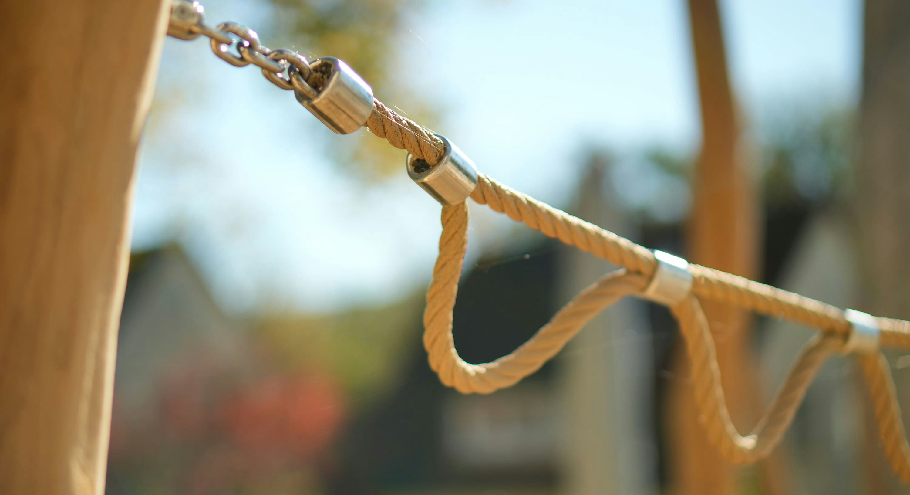Nahaufnahme eines beigen Seilzauns mit Metallverbindungen, der zwischen Holzpfosten gespannt ist. Häuser und Bäume sind im Hintergrund verschwommen.