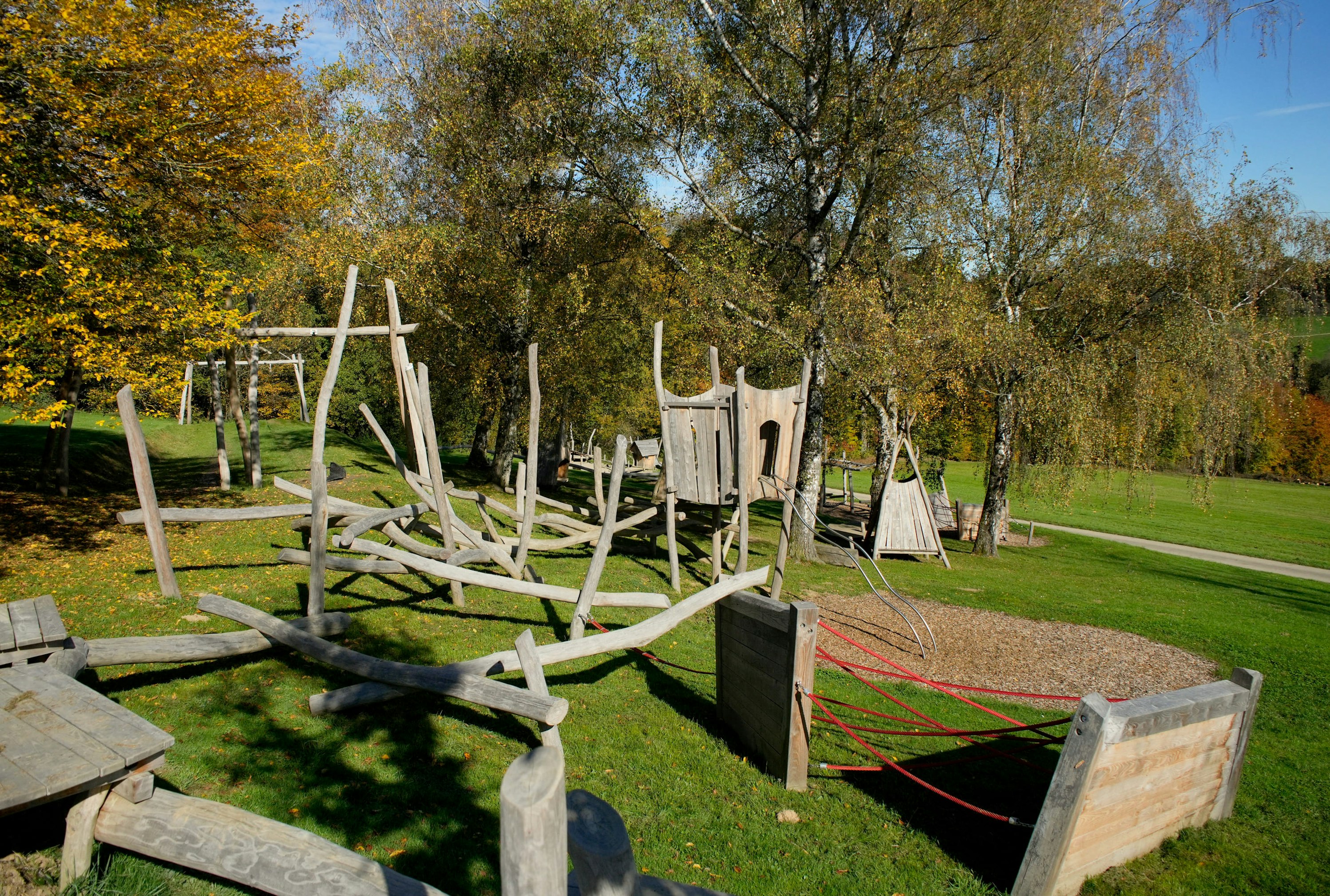 Eine hölzerne Spielplatzstruktur mit Seilen und Türmen steht in einem grasbewachsenen Park, umgeben von Herbstbäumen.
