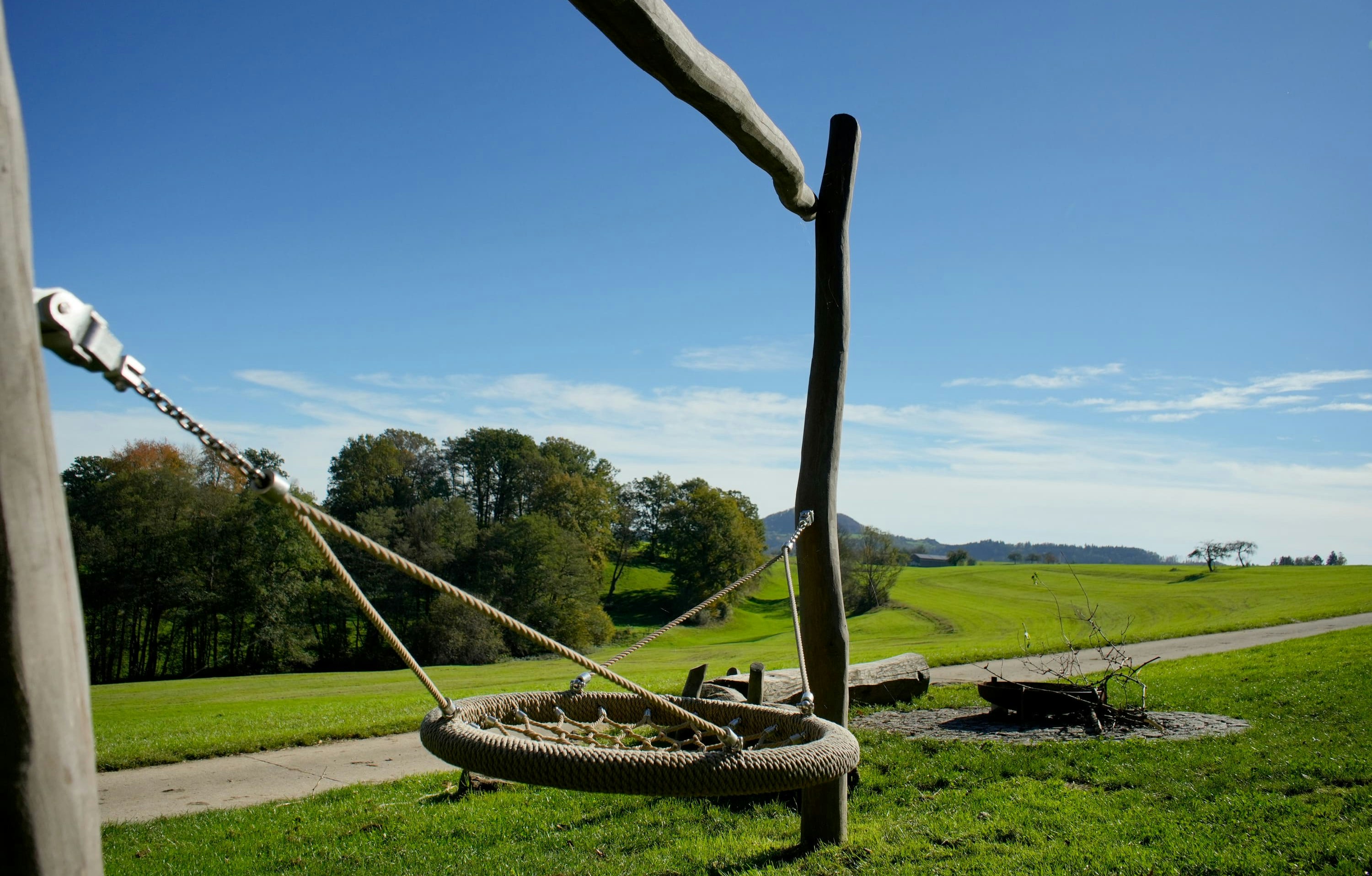 Eine Seilschaukel hängt an einem Holzrahmen auf einer Wiese. Unter einem klaren blauen Himmel sind ein Weg und Bäume zu sehen.