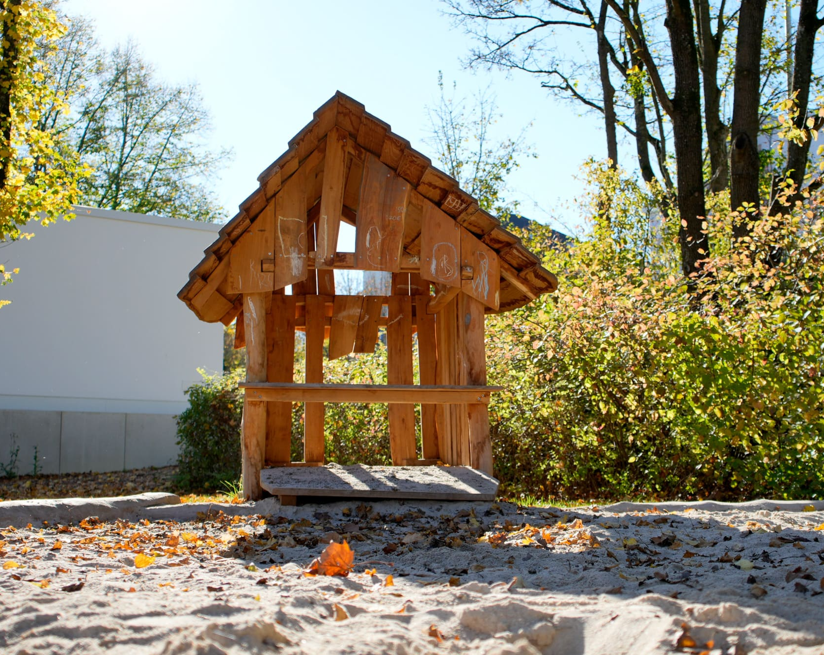 Holzspielhaus mit schrägem Dach auf einem Sandspielplatz, umgeben von Bäumen und Sträuchern unter klarem Himmel.