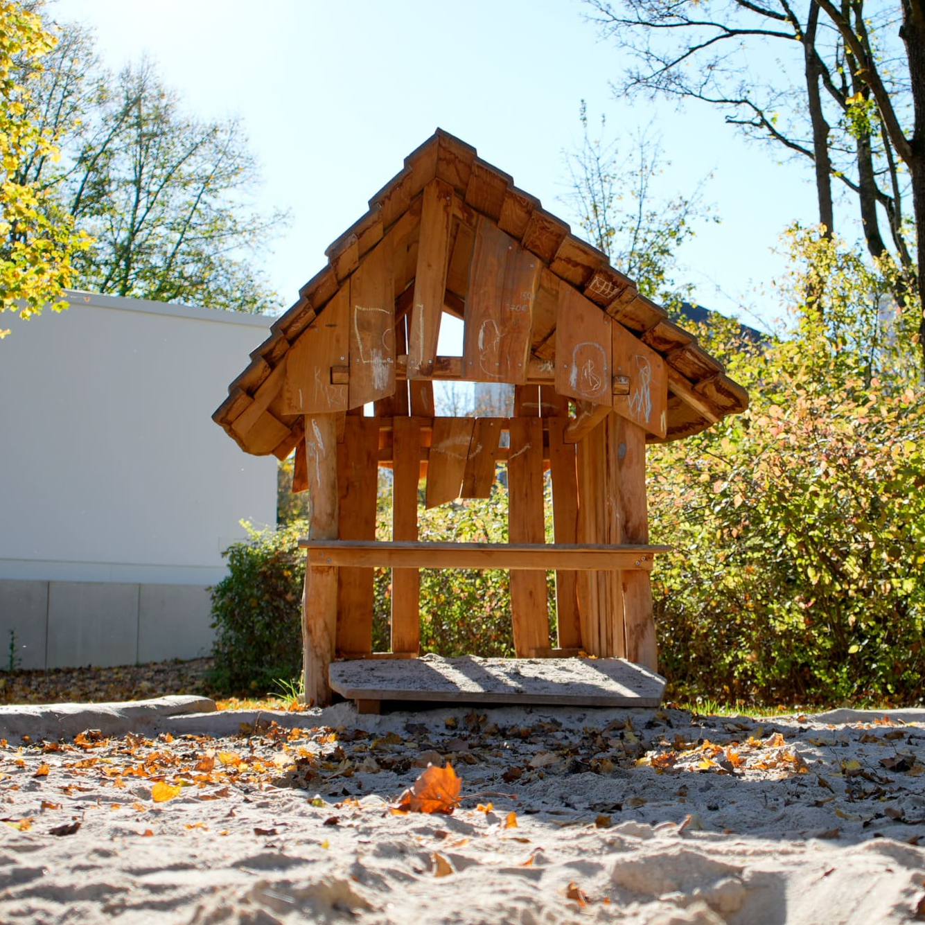 Holzspielhaus mit schrägem Dach auf einem Sandspielplatz, umgeben von Bäumen und Sträuchern unter klarem Himmel.