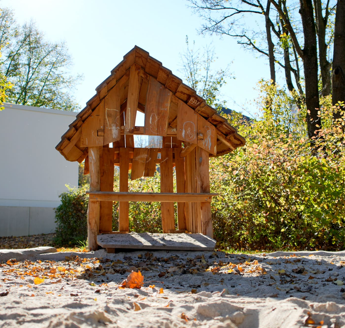 Holzspielhaus mit schrägem Dach auf einem Sandspielplatz, umgeben von Bäumen und Sträuchern unter klarem Himmel.