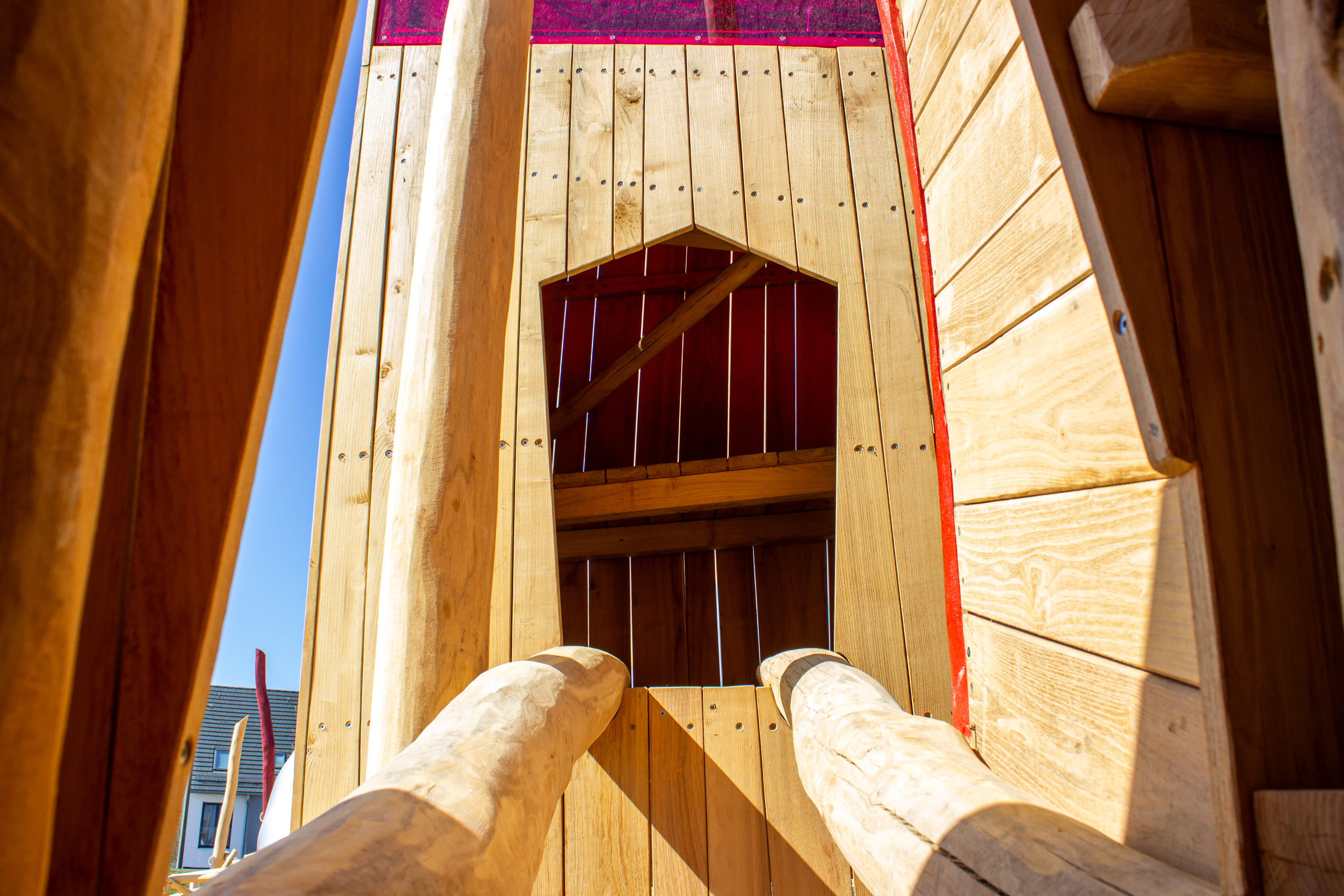 Hölzerne Spielplatzstruktur mit zwei abgewinkelten Baumstämmen, die zu einem Eingang führen. Helles Sonnenlicht wirft Schatten auf das Holz, und oben ist eine rote Platte sichtbar. Im Hintergrund blauer Himmel.
