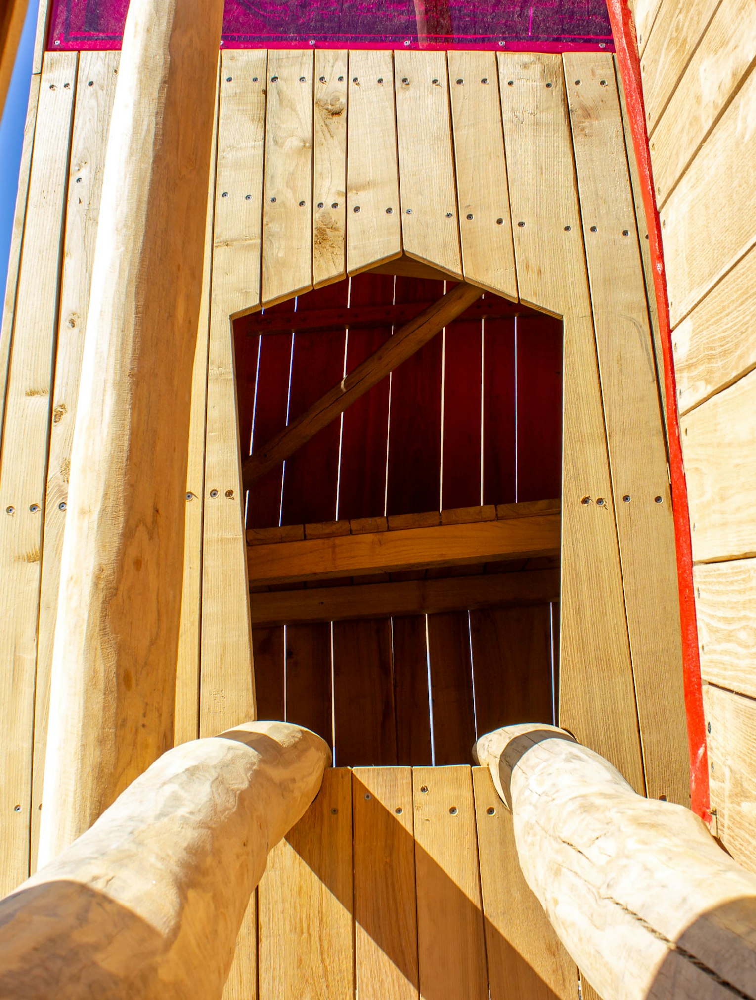 Hölzerne Spielplatzstruktur mit zwei abgewinkelten Baumstämmen, die zu einem Eingang führen. Helles Sonnenlicht wirft Schatten auf das Holz, und oben ist eine rote Platte sichtbar. Im Hintergrund blauer Himmel.