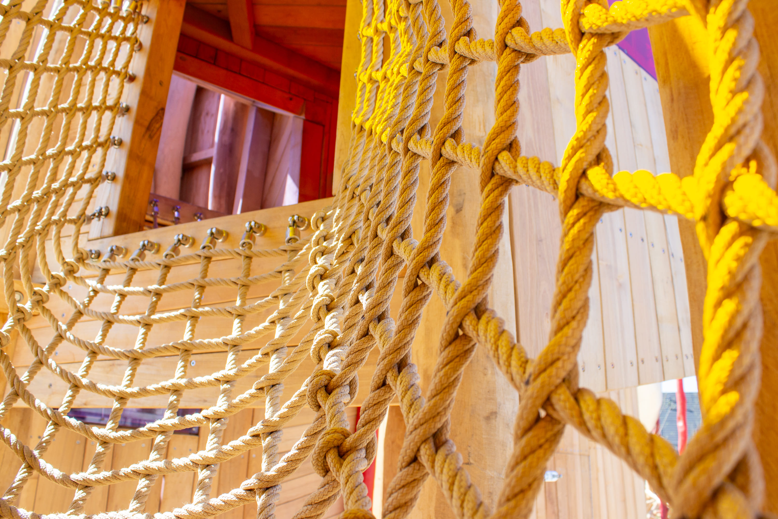 Nahaufnahme einer hölzernen Spielplatzstruktur mit ineinander verschlungenen Kletterseilen, die Knoten und Netzmuster vor einem Hintergrund aus Holzbrettern zeigen.