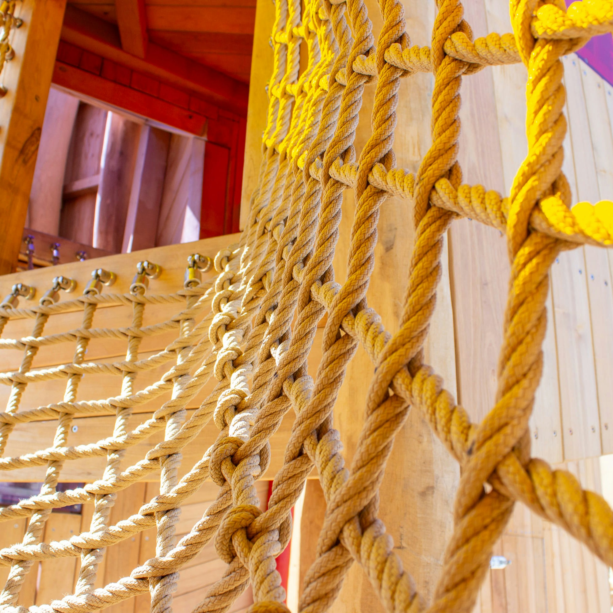 Nahaufnahme einer hölzernen Spielplatzstruktur mit ineinander verschlungenen Kletterseilen, die Knoten und Netzmuster vor einem Hintergrund aus Holzbrettern zeigen.
