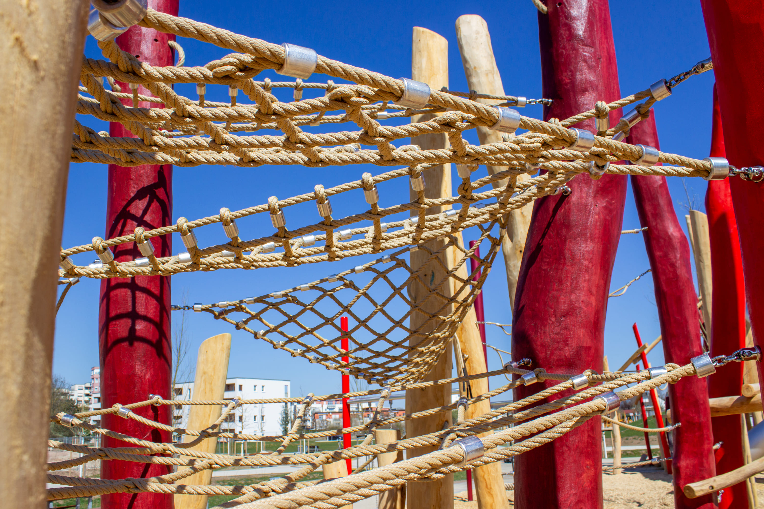Seilklettergerüst auf einem Spielplatz mit roten und natürlichen Holzstangen vor einem klaren blauen Himmel.