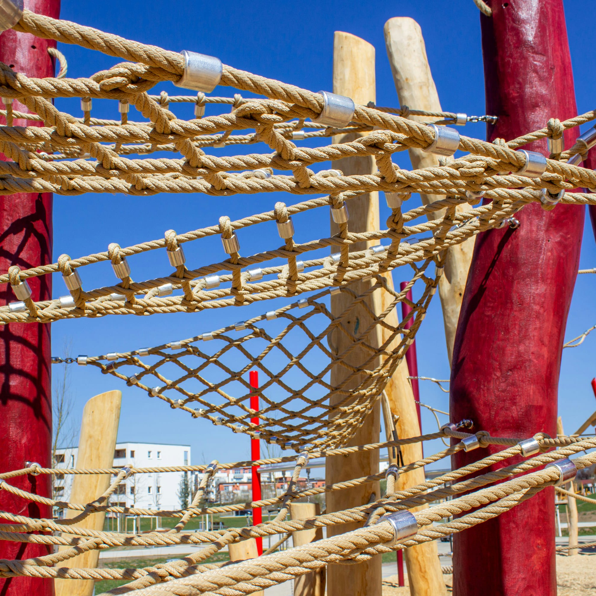 Seilklettergerüst auf einem Spielplatz mit roten und natürlichen Holzstangen vor einem klaren blauen Himmel.