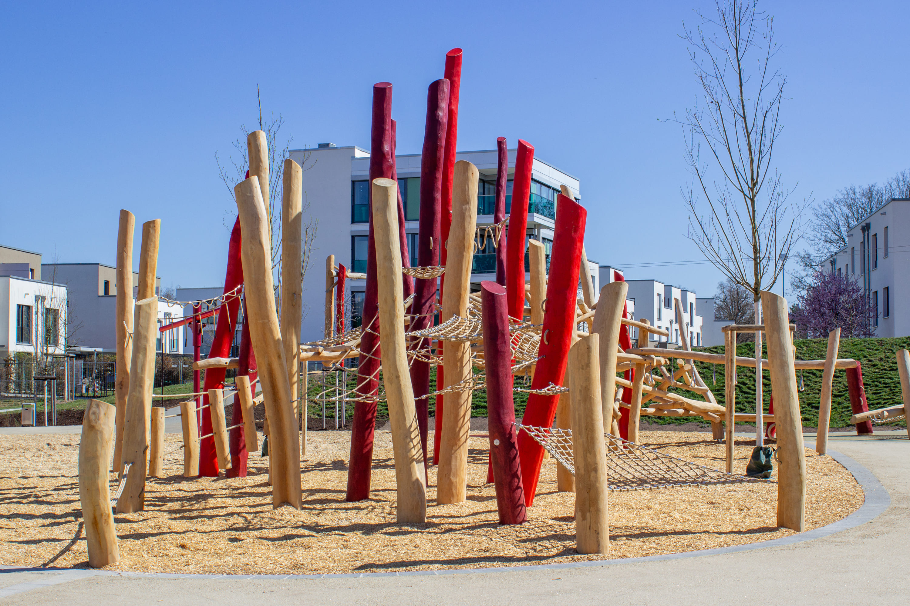 Spielplatz mit Holzkonstruktionen und roten Pfosten, die durch Seile miteinander verbunden sind, in einem Wohngebiet unter einem klaren blauen Himmel.