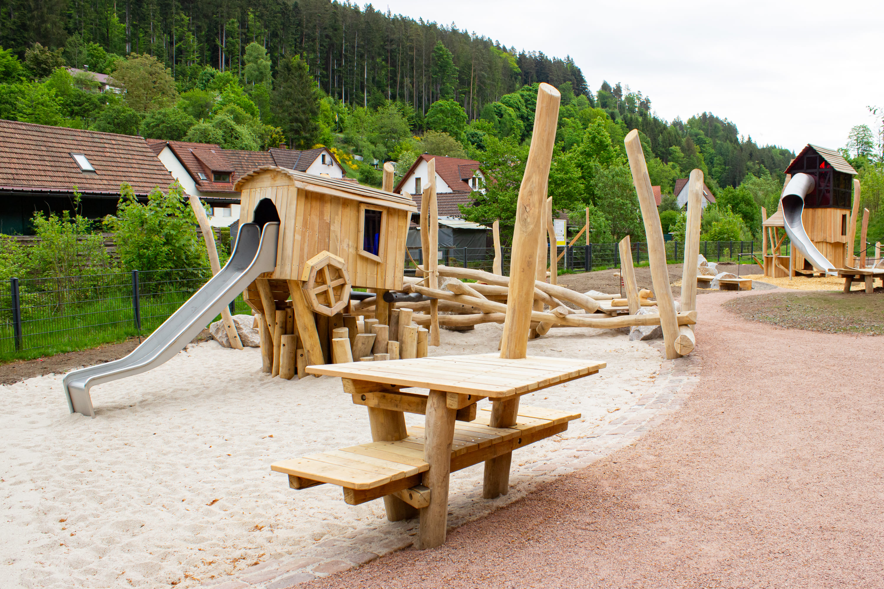 Holzspielplatzset mit Rutschen, Klettergerüsten und Sandbereich, neben einem hölzernen Picknicktisch, umgeben von Bäumen und Häusern im Hintergrund.
