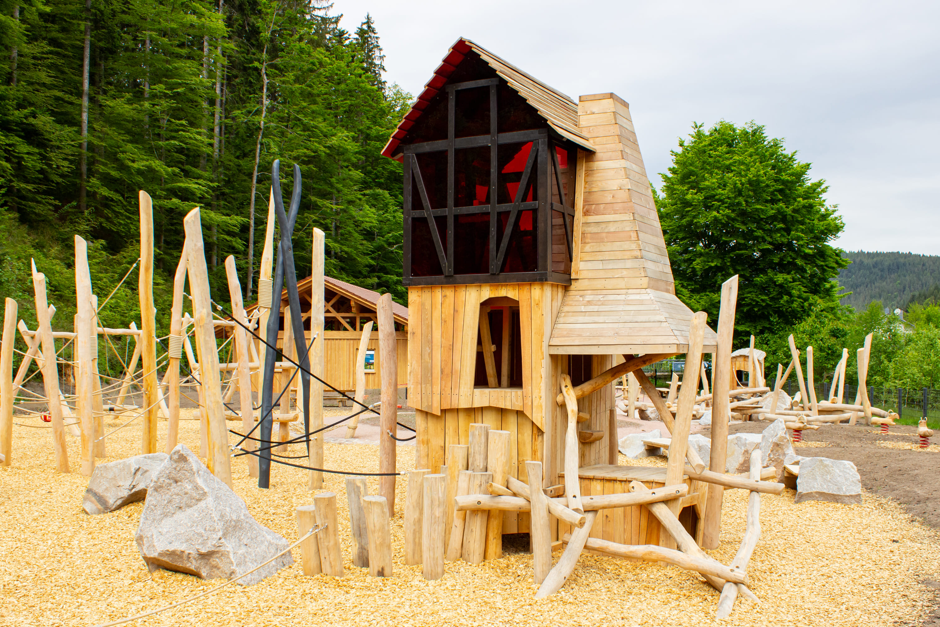 Hölzerne Spielplatzstruktur mit Baumstämmen, Seilen, Felsen und einem zentralen Turm, umgeben von Bäumen und Holzspänen unter einem bewölkten Himmel.