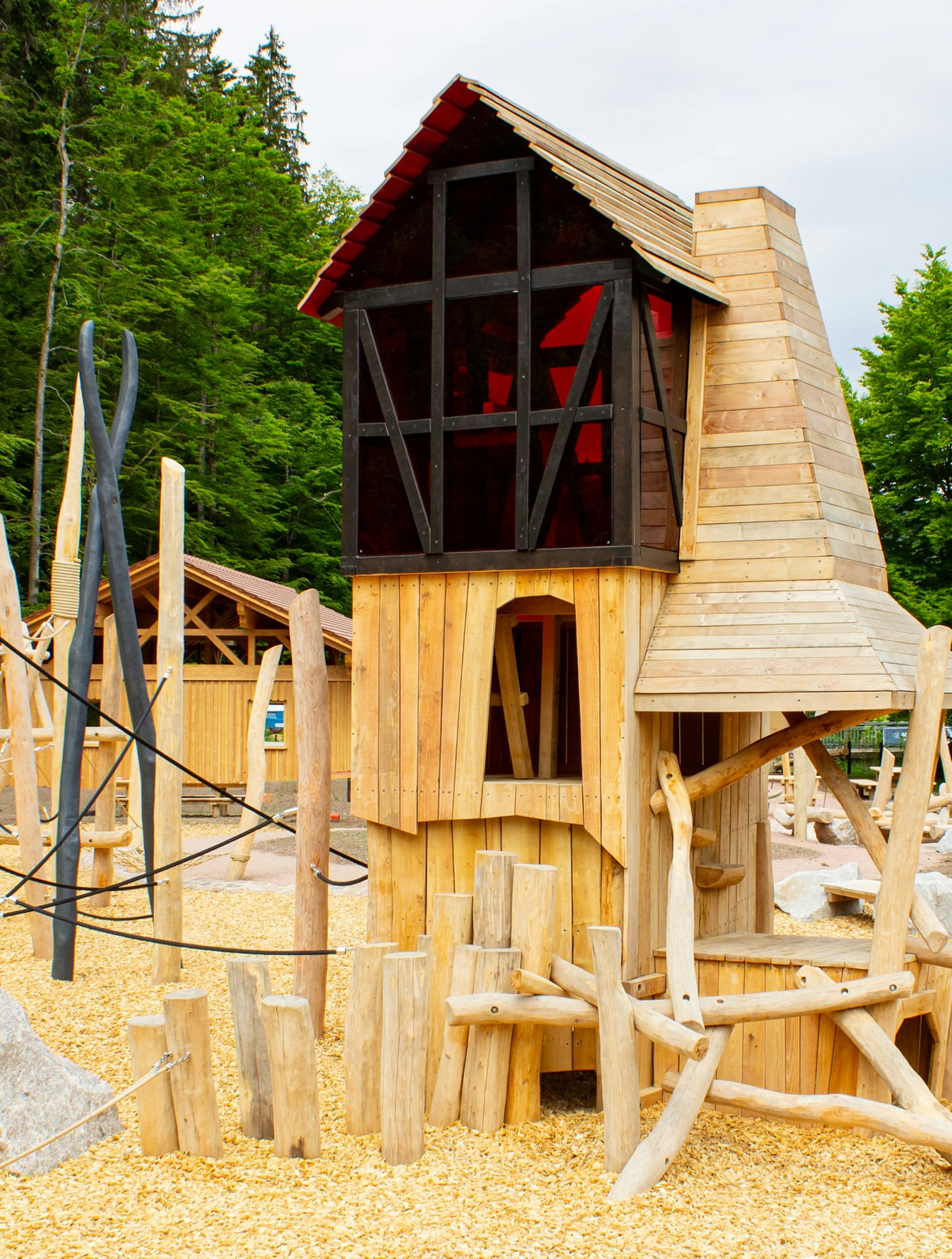 Hölzerne Spielplatzstruktur mit Baumstämmen, Seilen, Felsen und einem zentralen Turm, umgeben von Bäumen und Holzspänen unter einem bewölkten Himmel.
