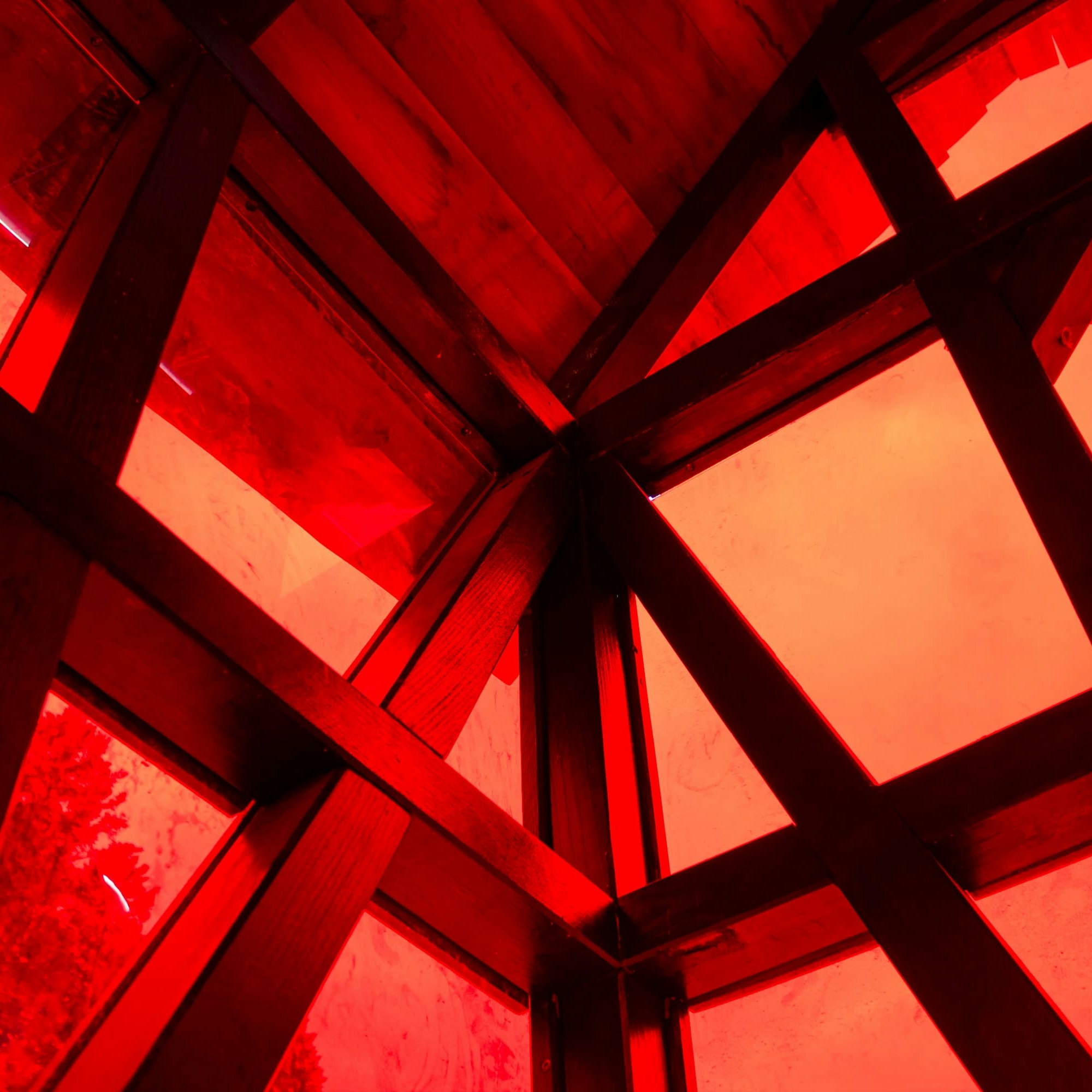 Holzbalken kreuzen sich mit rot getönten Platten in einer geometrischen Deckenstruktur und erzeugen ein kräftiges Muster aus Linien und Formen.