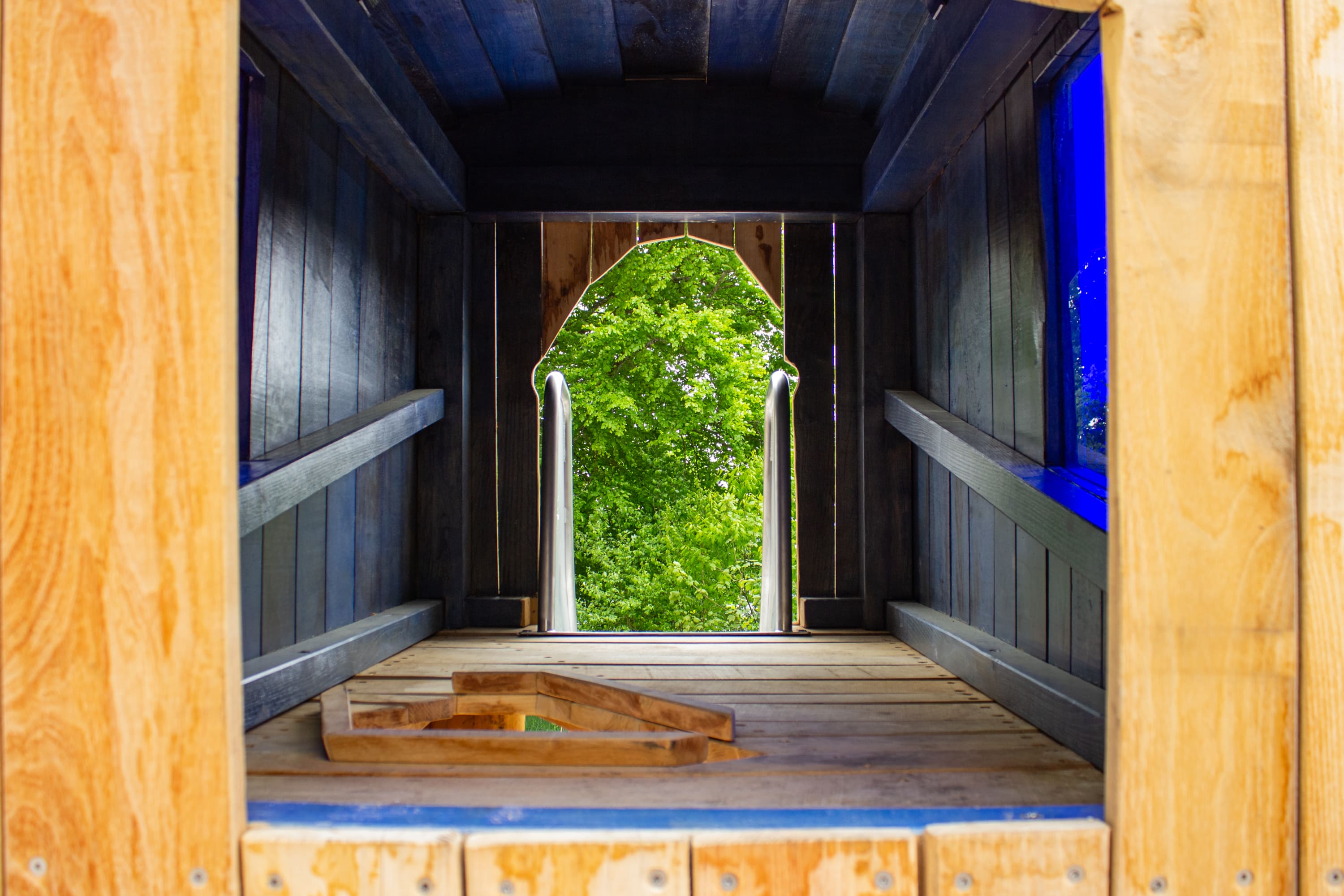Innenansicht einer hölzernen Spielplatz-Tunnelrutsche mit einem gewölbten Ausgang, der zu den grünen Bäumen draußen führt.