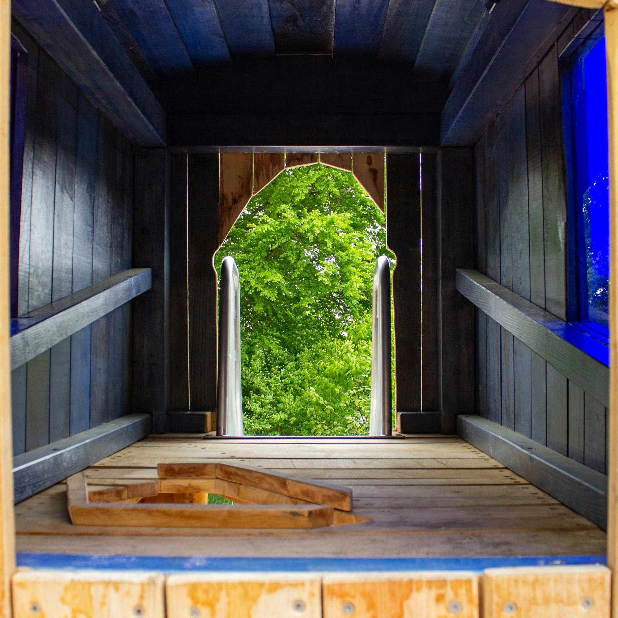 Innenansicht einer hölzernen Spielplatz-Tunnelrutsche mit einem gewölbten Ausgang, der zu den grünen Bäumen draußen führt.