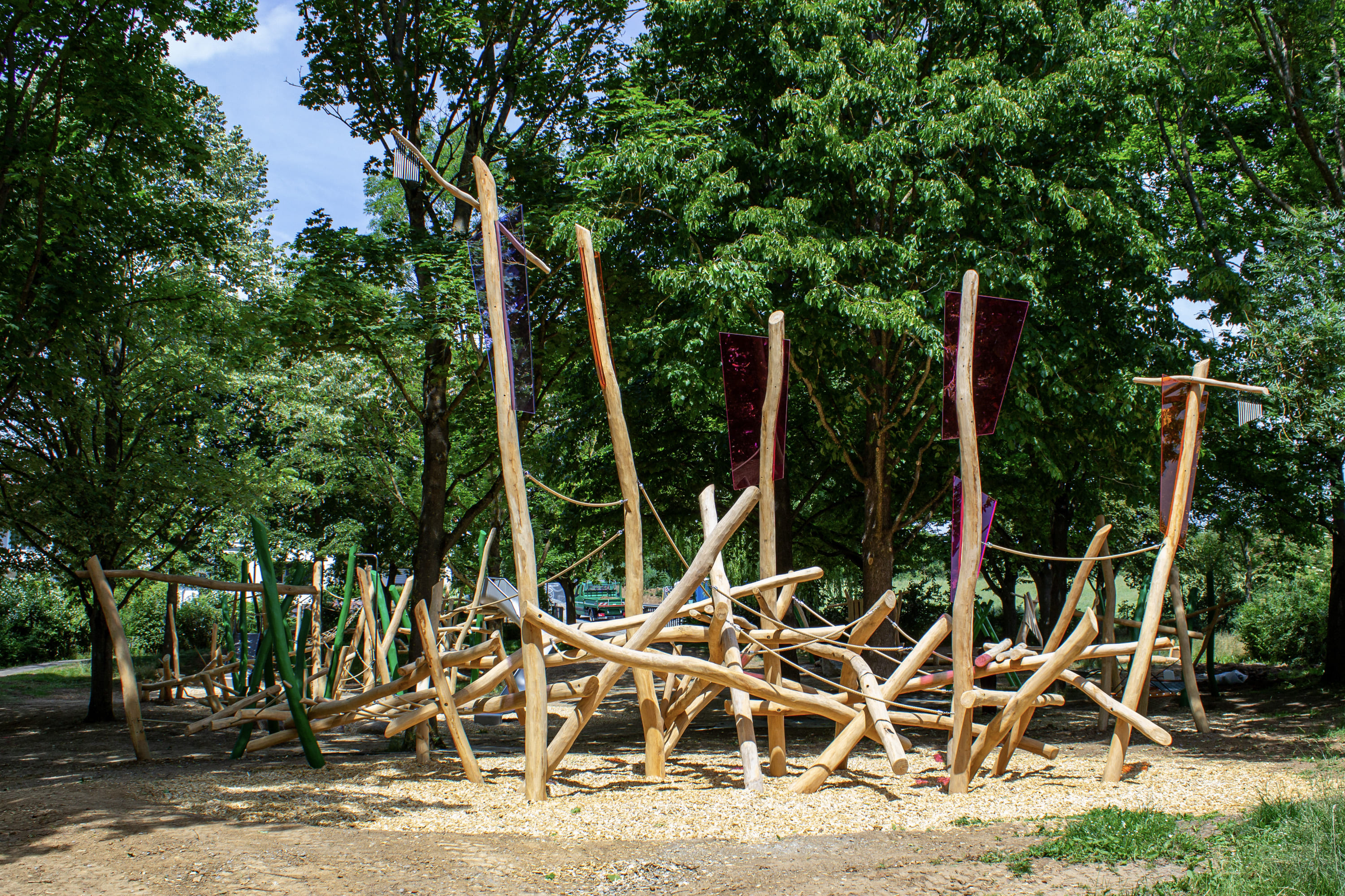Ein hölzernes Spielgerät aus unregelmäßigen Stämmen und Seilen steht zwischen Bäumen auf einer gemulchten Fläche in einem Park.