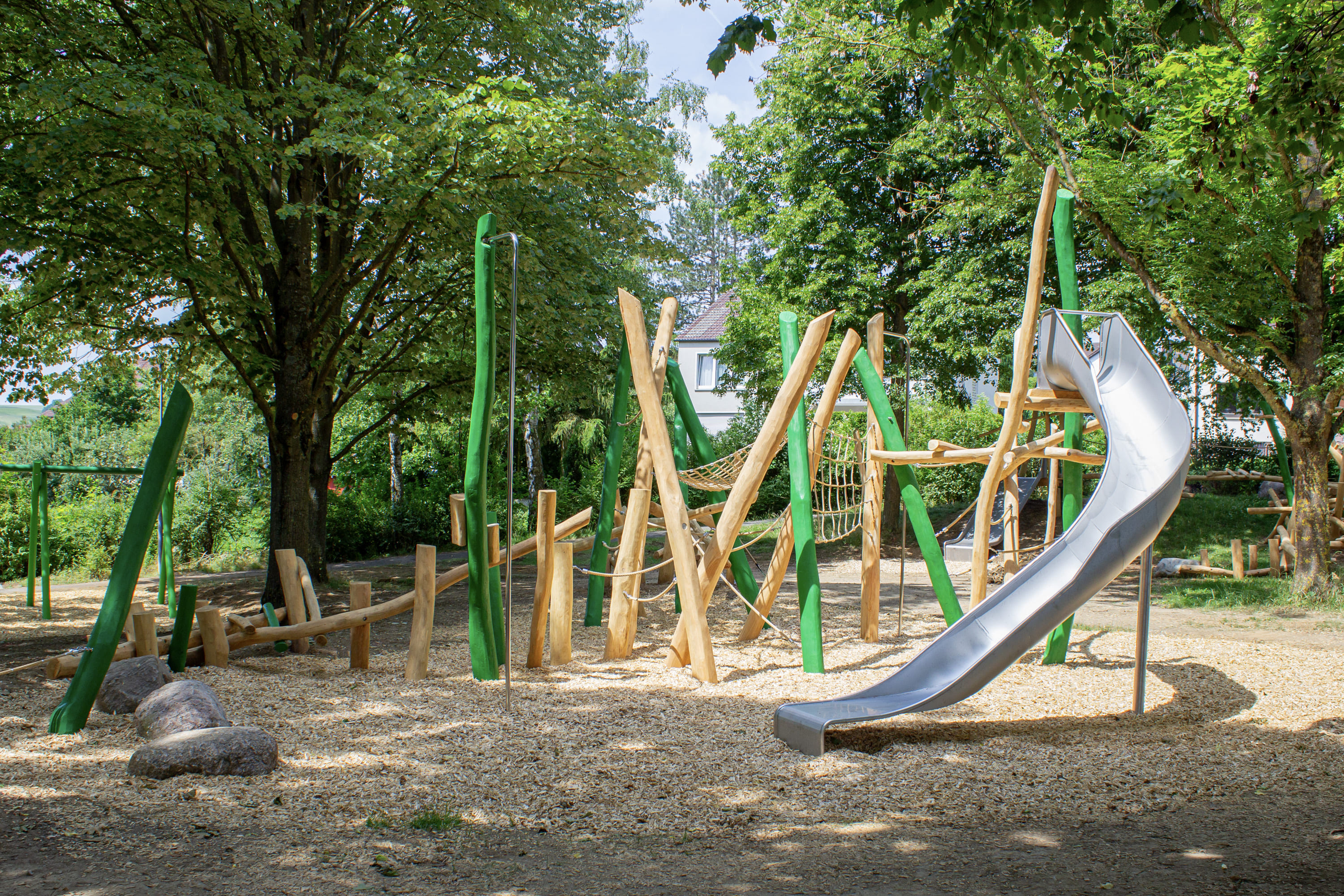 Spielplatz im Freien mit Klettergerüsten aus Holz, einer Netzbrücke und einer Metallrutsche, umgeben von Bäumen und Holzspänen auf dem Boden.