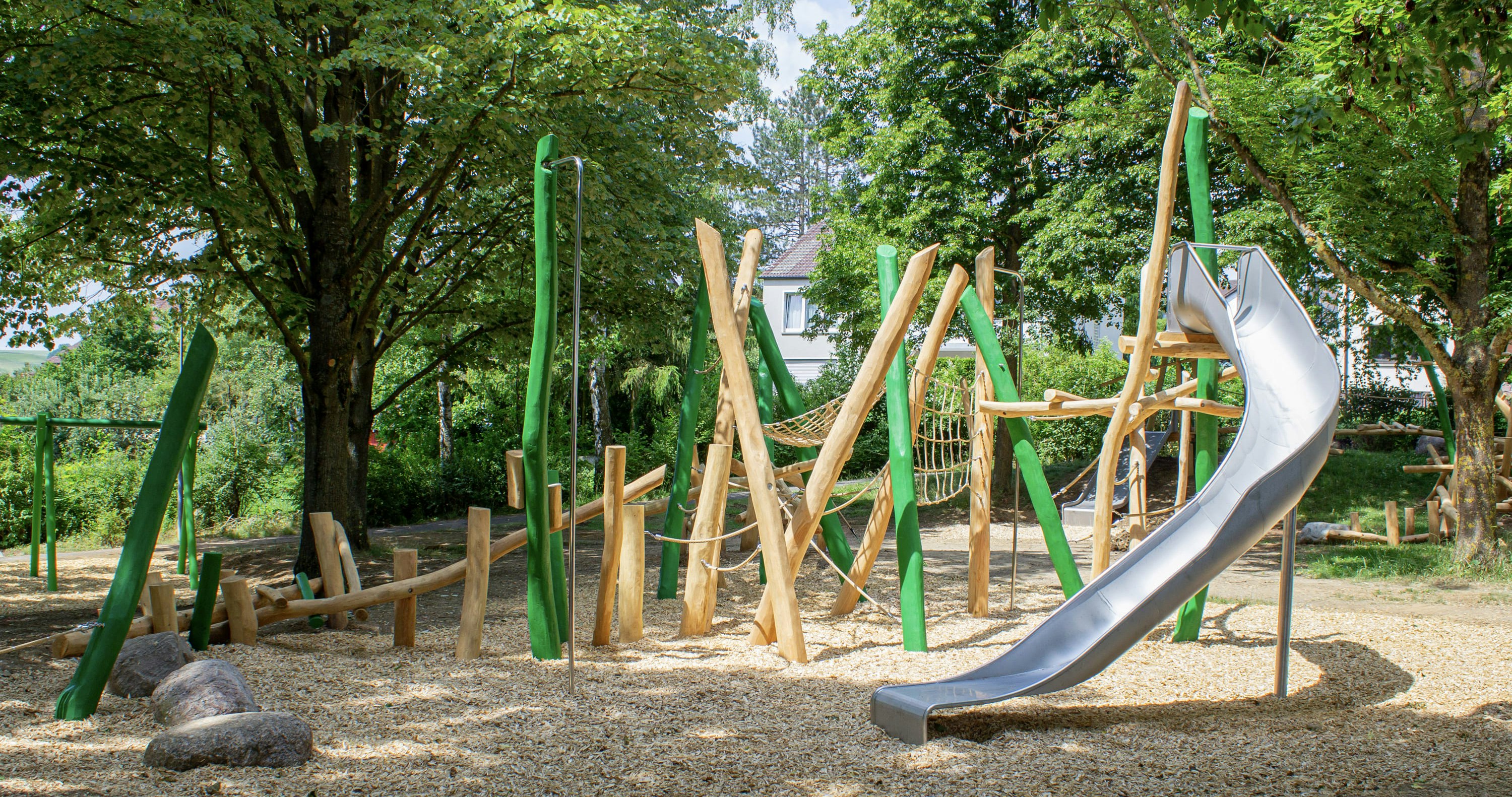 Spielplatz im Freien mit Klettergerüsten aus Holz, einer Netzbrücke und einer Metallrutsche, umgeben von Bäumen und Holzspänen auf dem Boden.