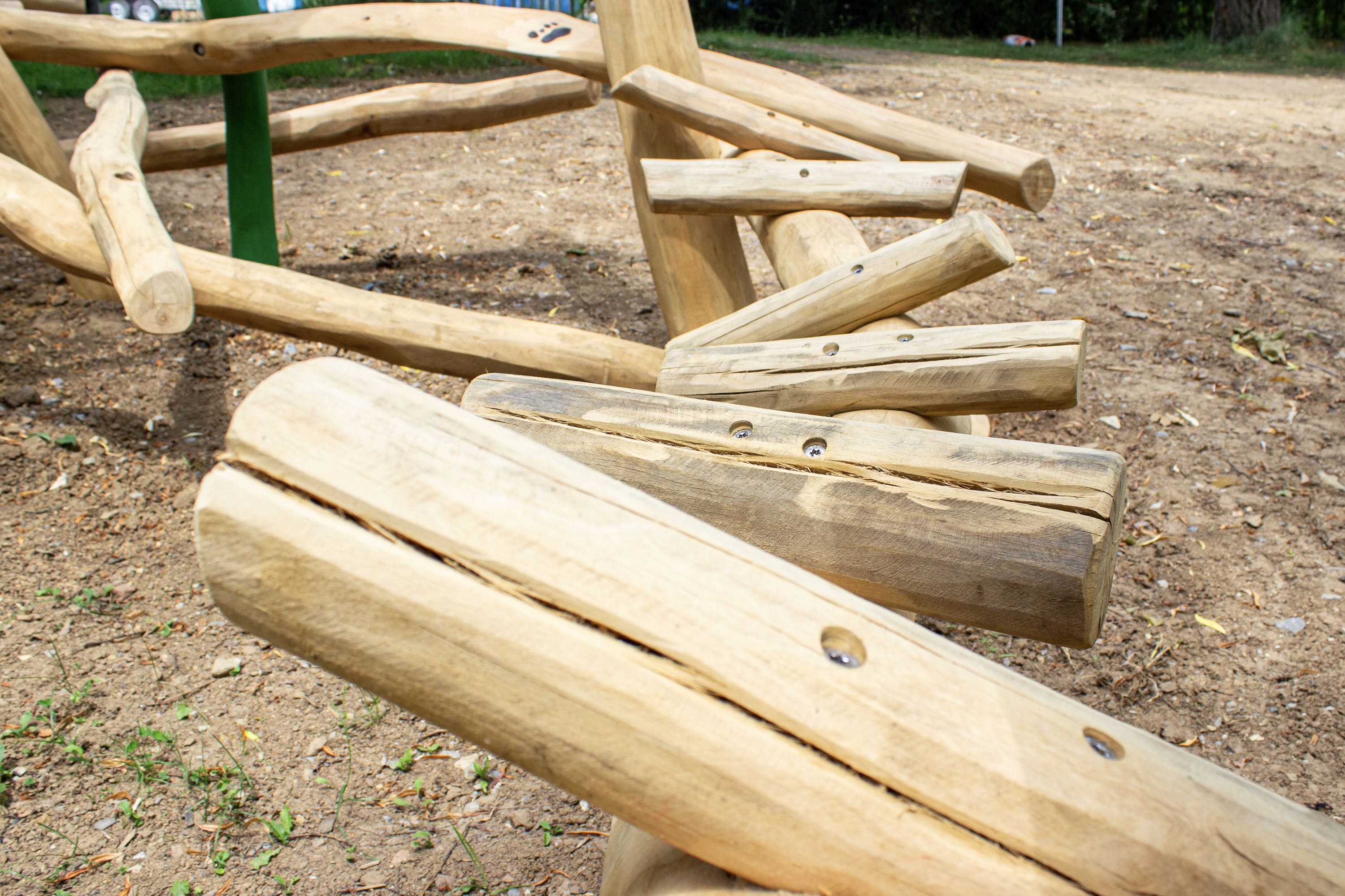 Nahaufnahme eines Spielplatzgeräts aus Holz, das aus miteinander verbundenen Naturstämmen besteht und auf einem unbefestigten Boden in einem Außenbereich aufgestellt ist.