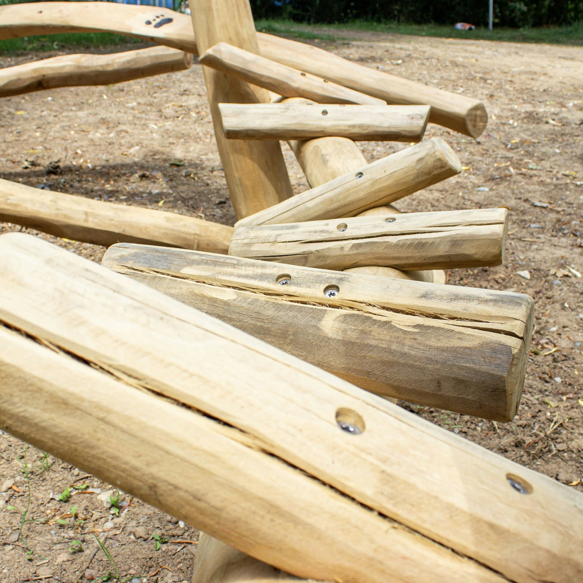 Nahaufnahme eines Spielplatzgeräts aus Holz, das aus miteinander verbundenen Naturstämmen besteht und auf einem unbefestigten Boden in einem Außenbereich aufgestellt ist.