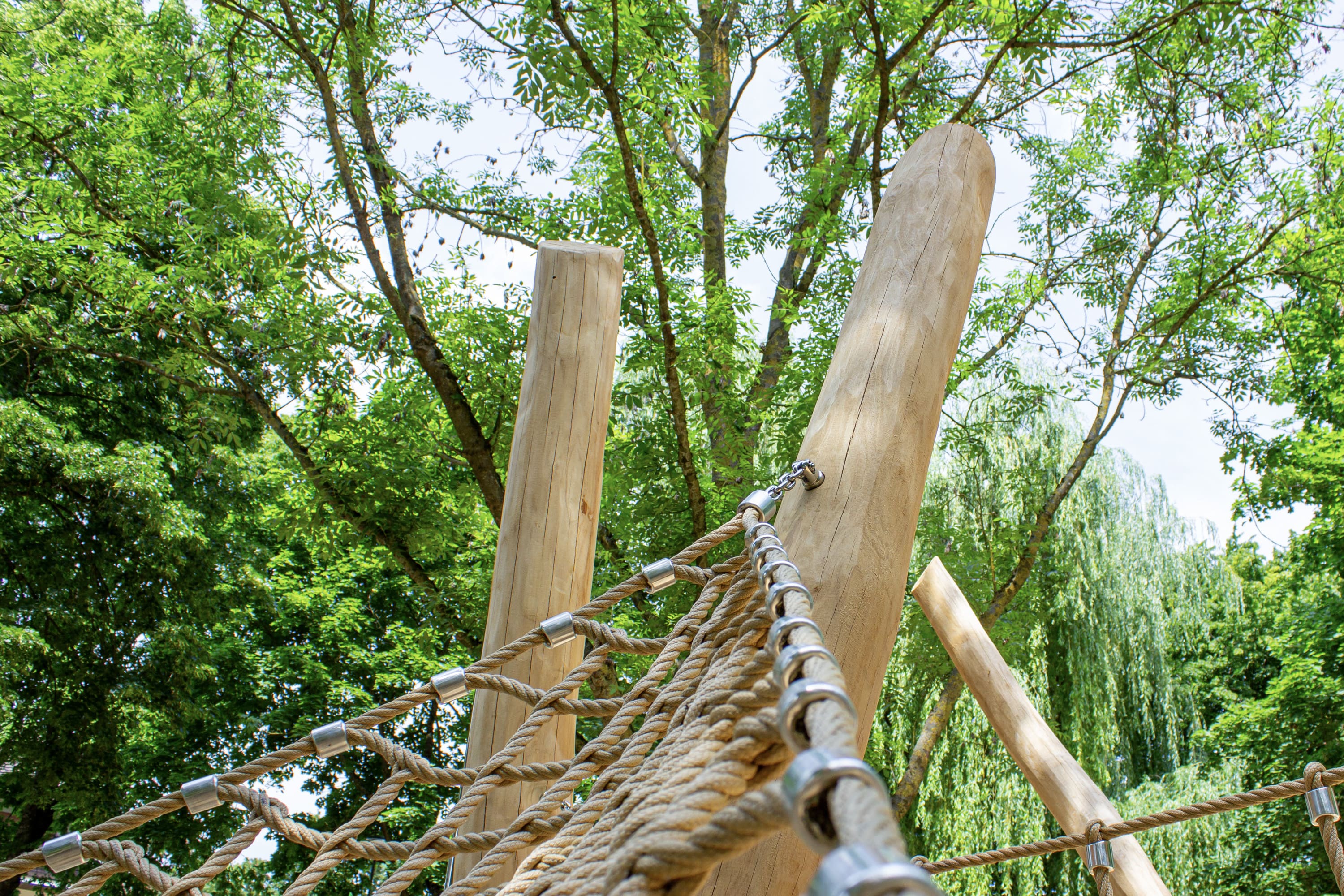 An hohen Holzpfosten befestigtes Seilkletternetz auf einem Spielplatz im Freien, umgeben von grünen Bäumen und Blättern.