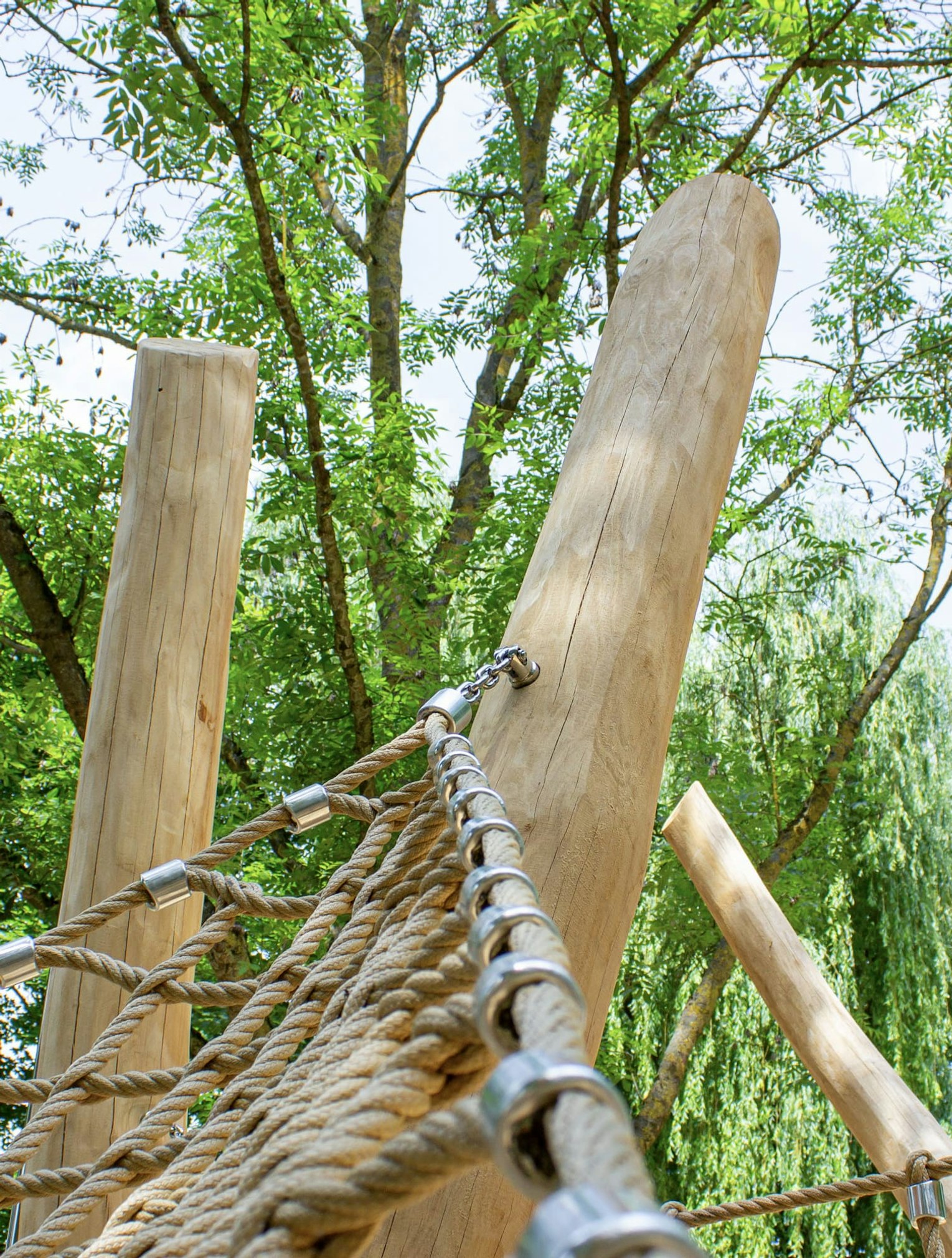 An hohen Holzpfosten befestigtes Seilkletternetz auf einem Spielplatz im Freien, umgeben von grünen Bäumen und Blättern.