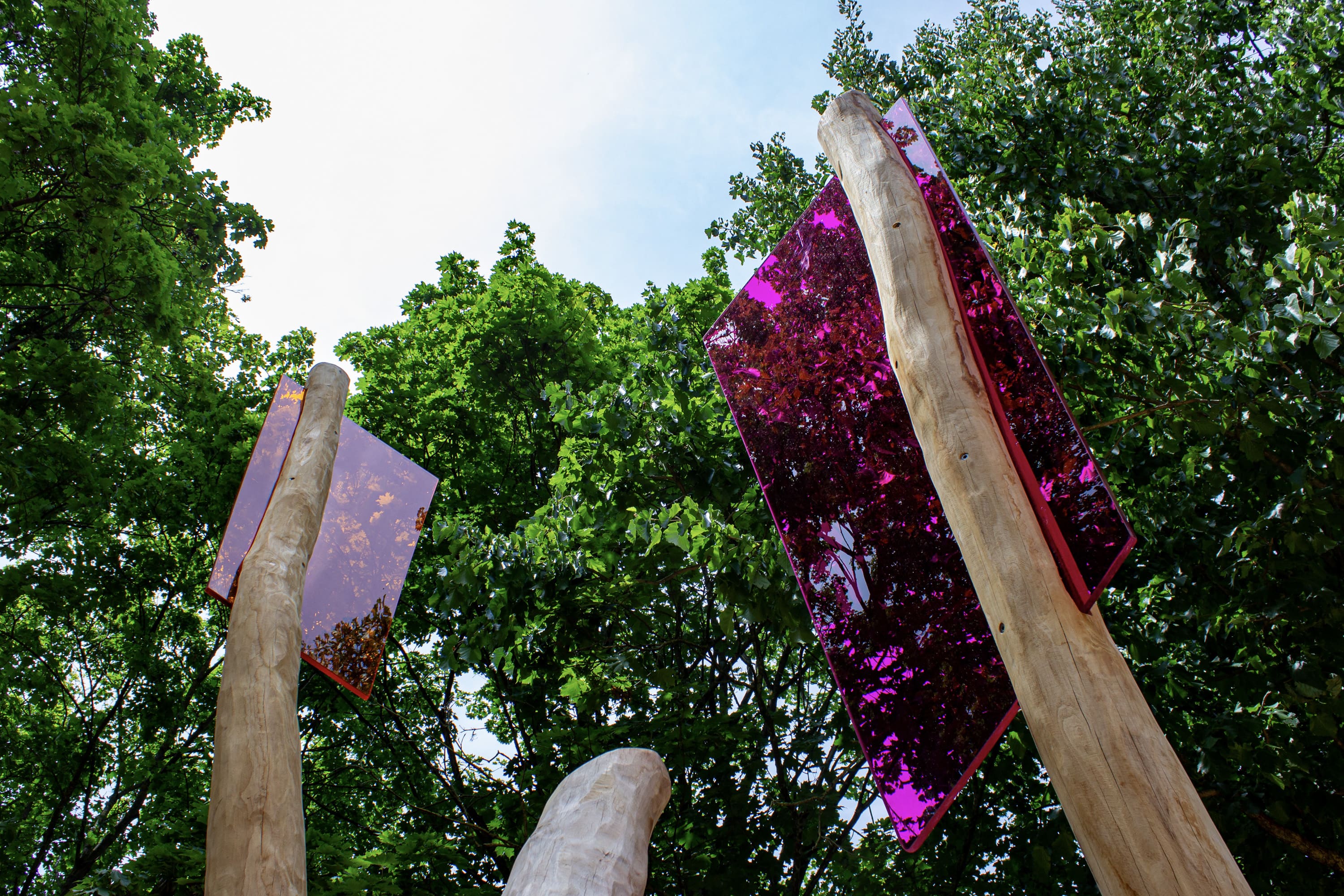Drei hohe Holzpfähle mit großen, reflektierenden rosafarbenen Scheiben an der Spitze, vor einem Hintergrund aus grünen Bäumen und blauem Himmel.