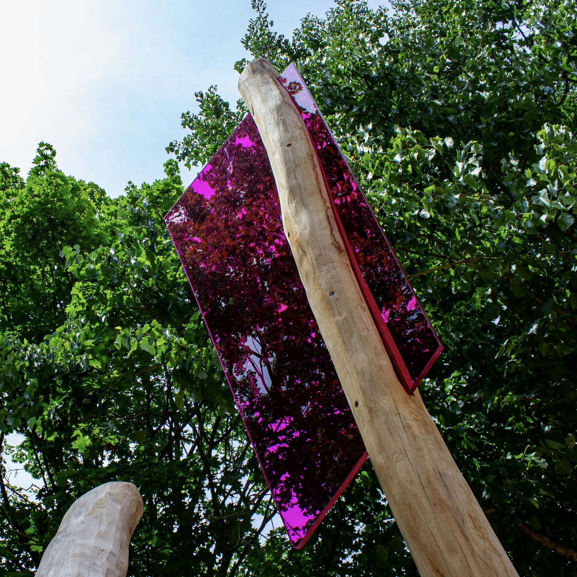 Drei hohe Holzpfähle mit großen, reflektierenden rosafarbenen Scheiben an der Spitze, vor einem Hintergrund aus grünen Bäumen und blauem Himmel.