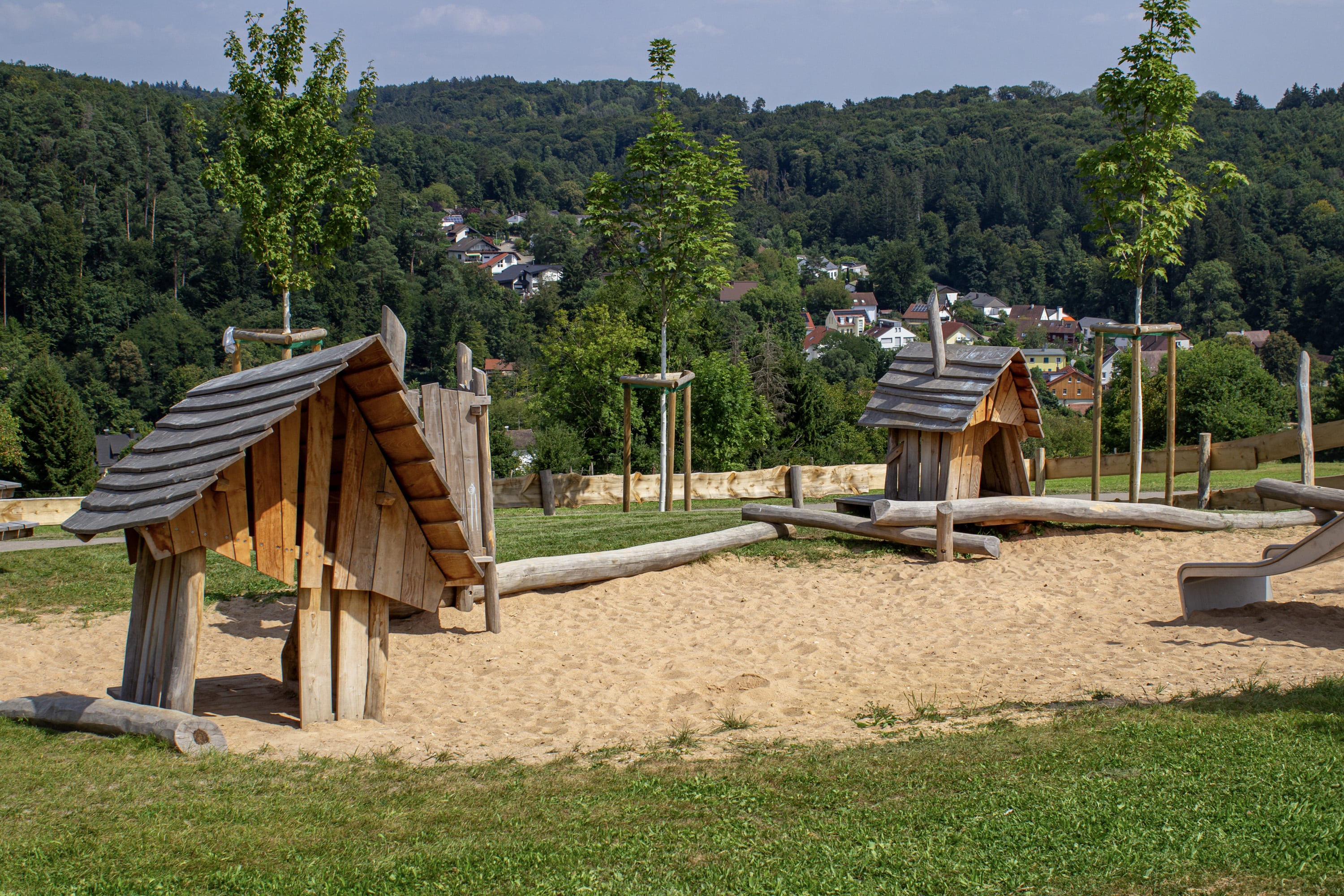 Die hölzernen Spielgeräte in Form kleiner Häuser stehen auf Sand, umgeben von Gras und jungen Bäumen, mit einem bewaldeten Hügel und Häusern im Hintergrund.