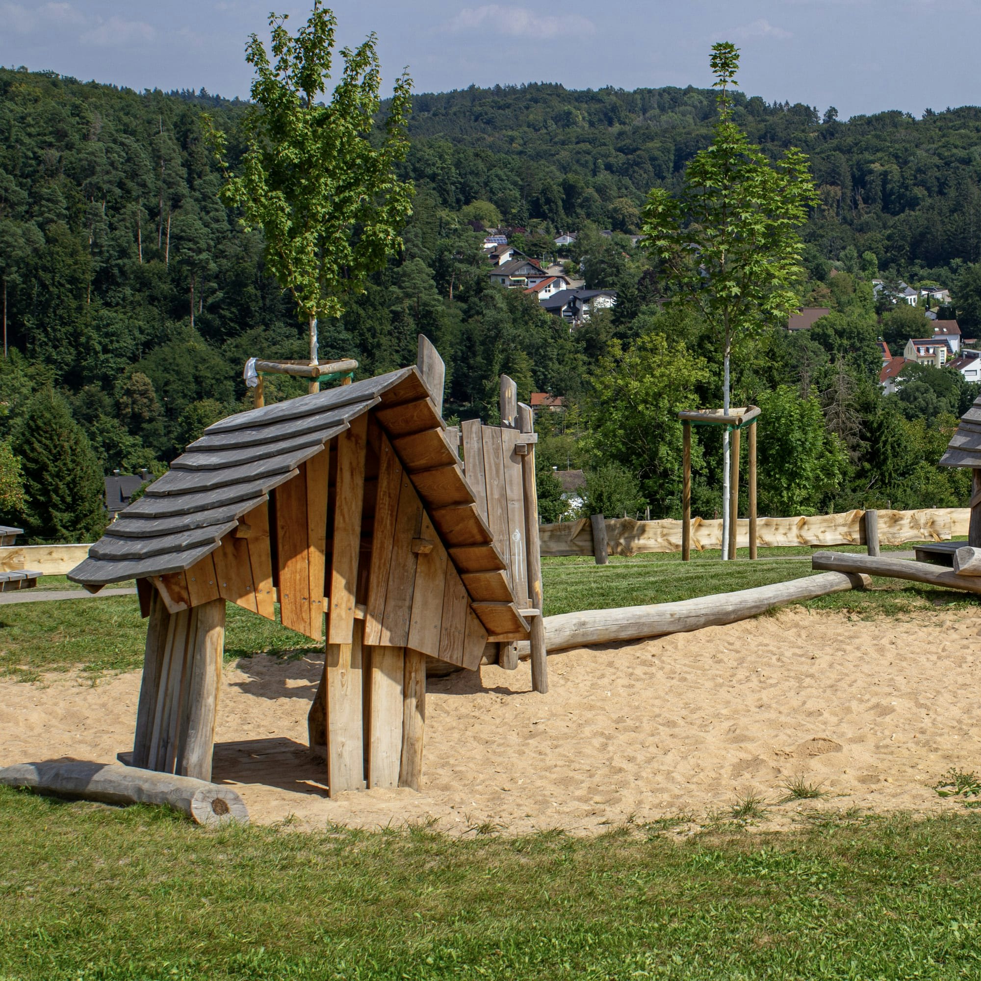Die hölzernen Spielgeräte in Form kleiner Häuser stehen auf Sand, umgeben von Gras und jungen Bäumen, mit einem bewaldeten Hügel und Häusern im Hintergrund.