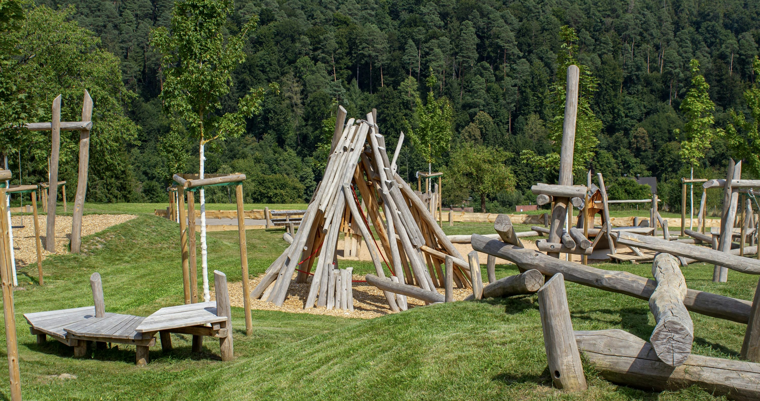 Ein natürlicher Spielplatz mit hölzernen Klettergerüsten und Bänken auf grasbewachsenem Gelände, mit einem Wald im Hintergrund.