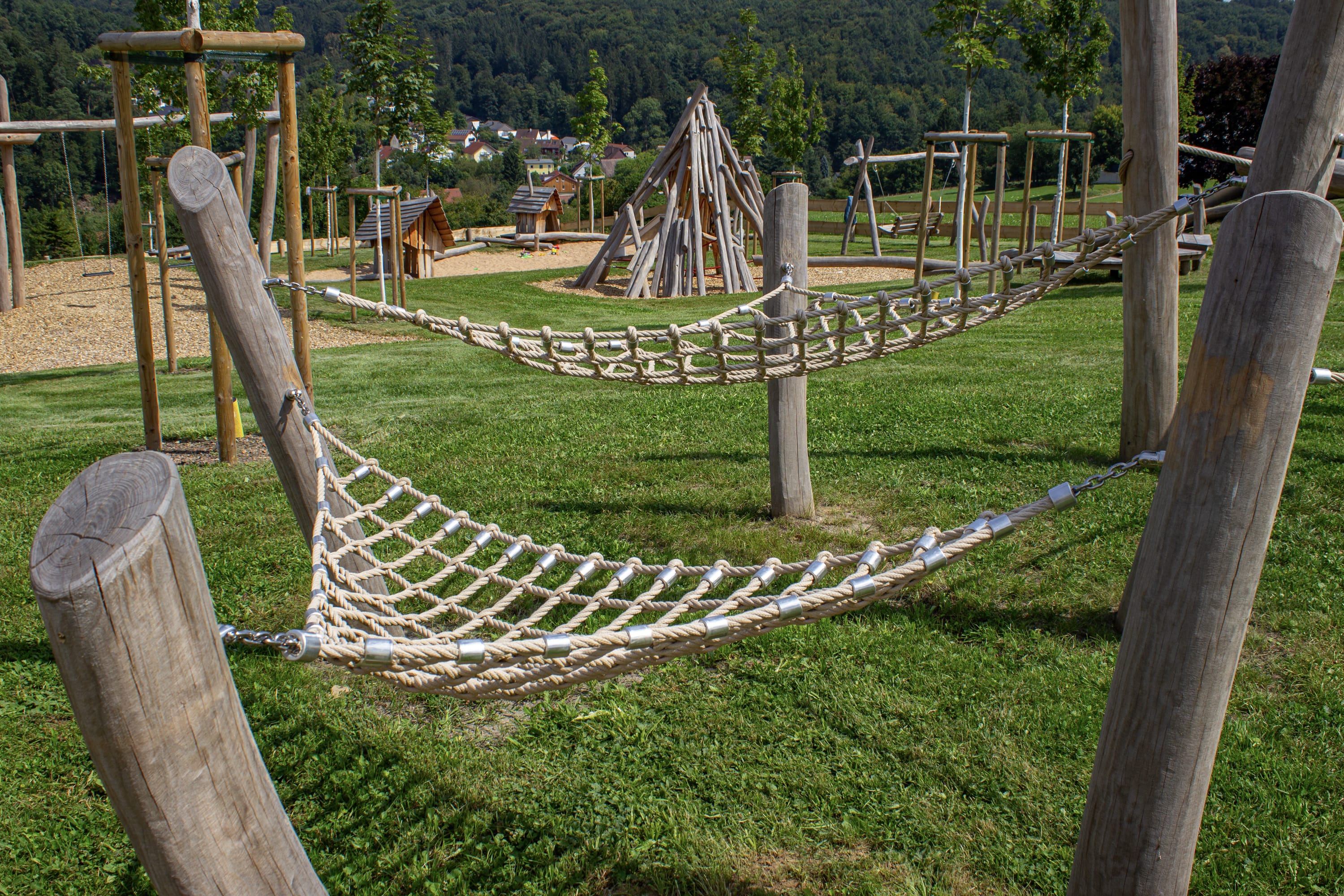 Zwei Seilhängematten sind zwischen Holzpfosten auf einem grasbewachsenen Spielplatz aufgehängt; im Hintergrund sind weitere Holzspielgeräte zu sehen.