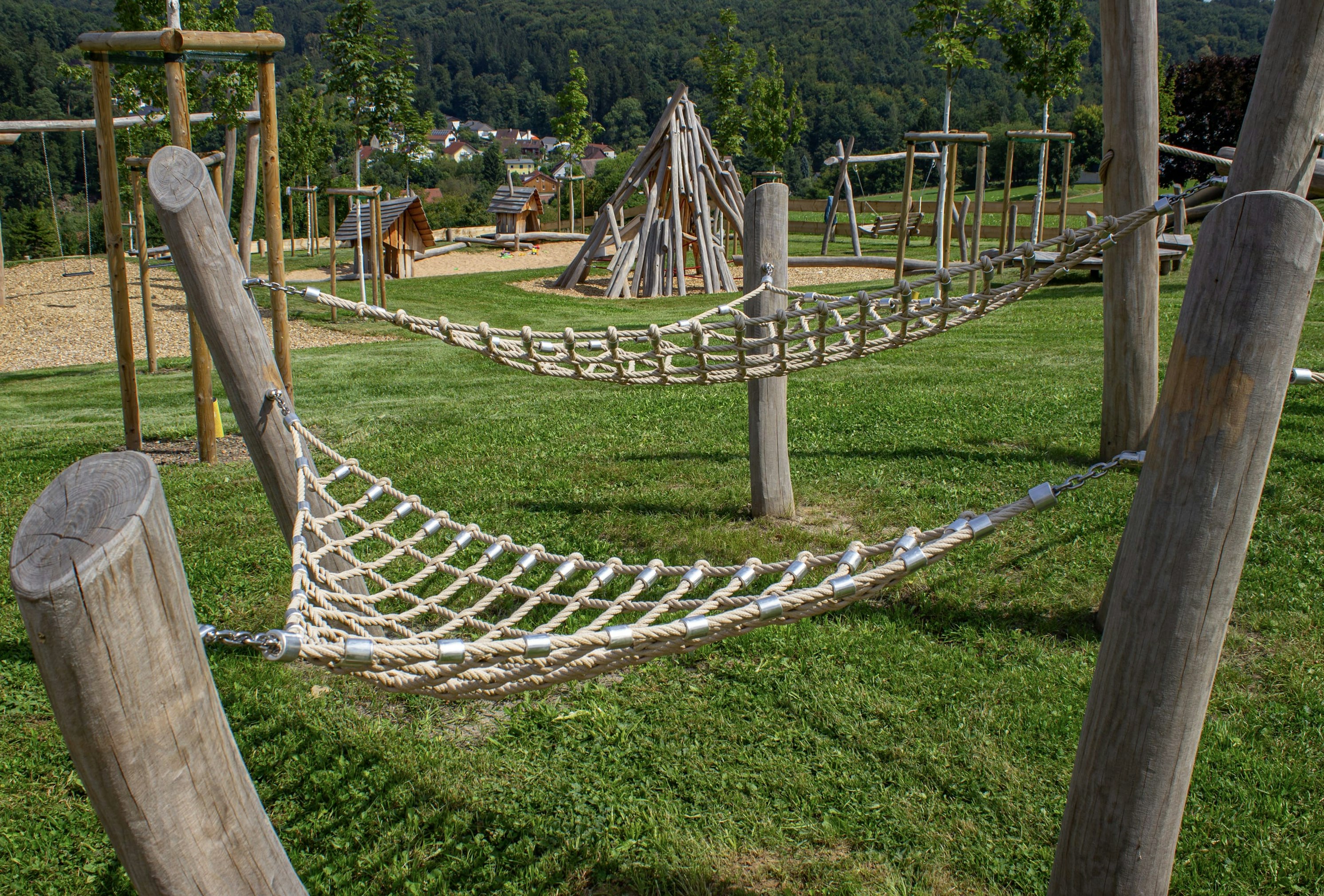 Zwei Seilhängematten sind zwischen Holzpfosten auf einem grasbewachsenen Spielplatz aufgehängt; im Hintergrund sind weitere Holzspielgeräte zu sehen.