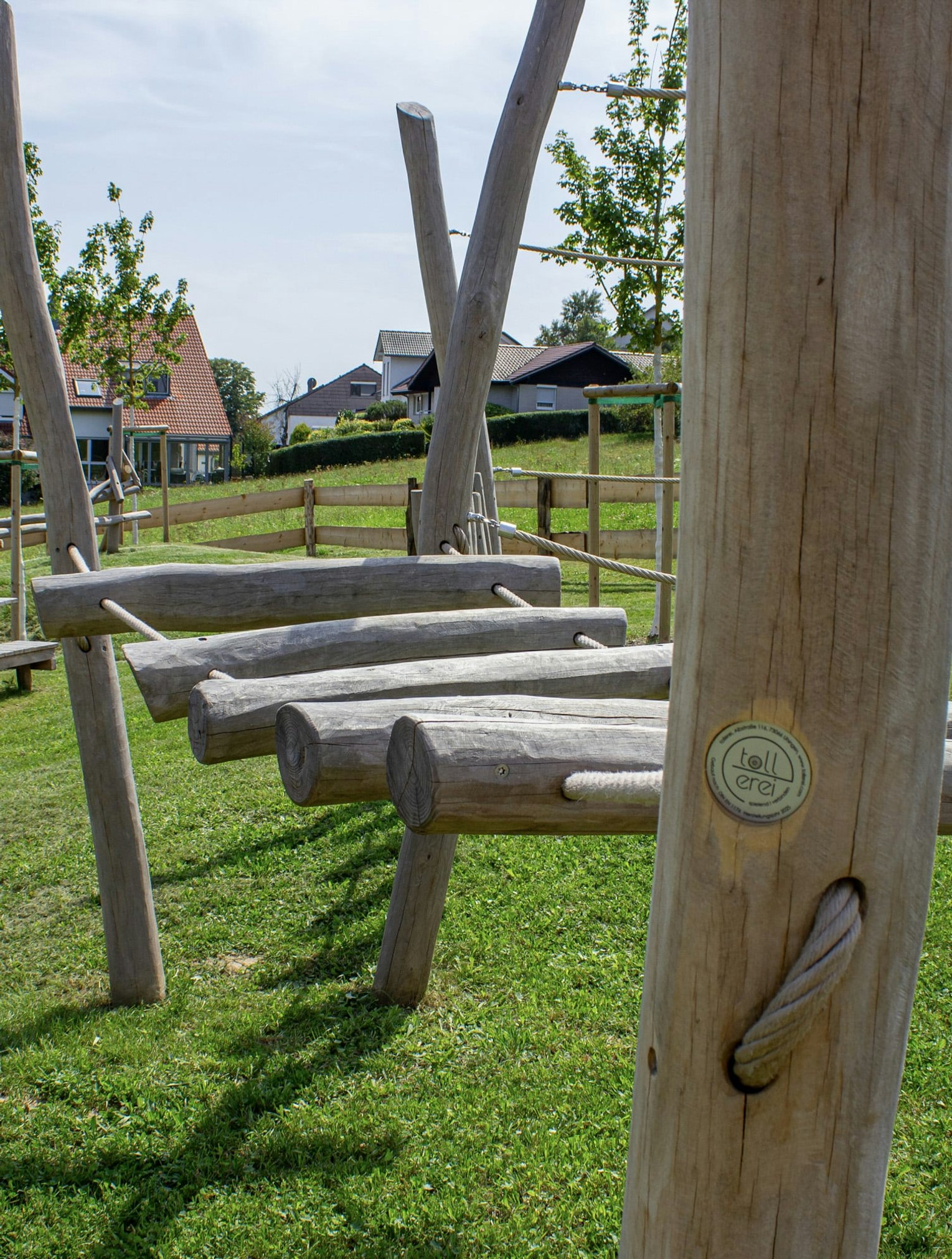 Spielplatz im Freien mit Klettergerüsten aus Holz und Picknicktischen auf einer Wiese, mit Häusern und Bäumen im Hintergrund.