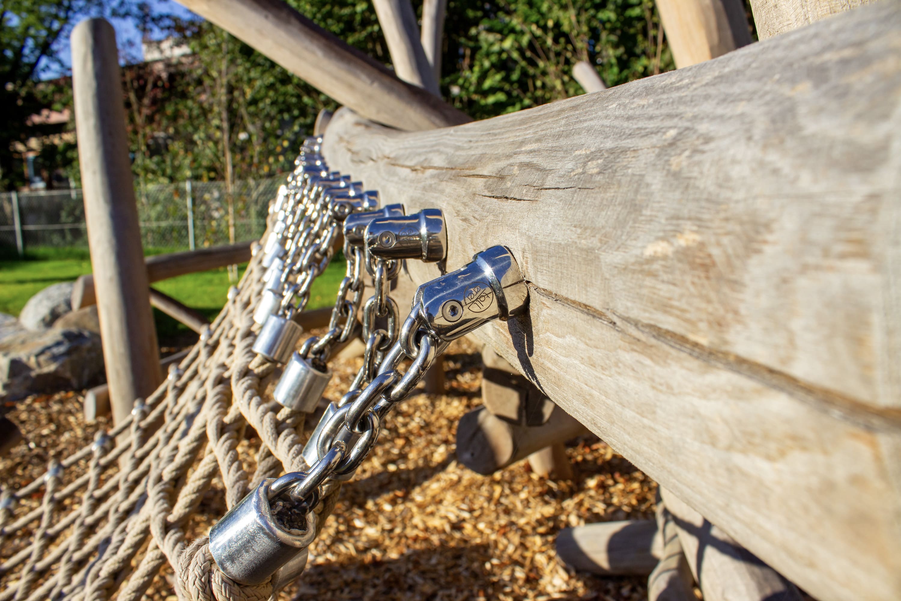 Nahaufnahme von Metallketten, die an einer Holzstruktur befestigt sind, die Teil eines Kletternetzes auf einem Spielplatz ist, mit Holzspänen auf dem Boden.
