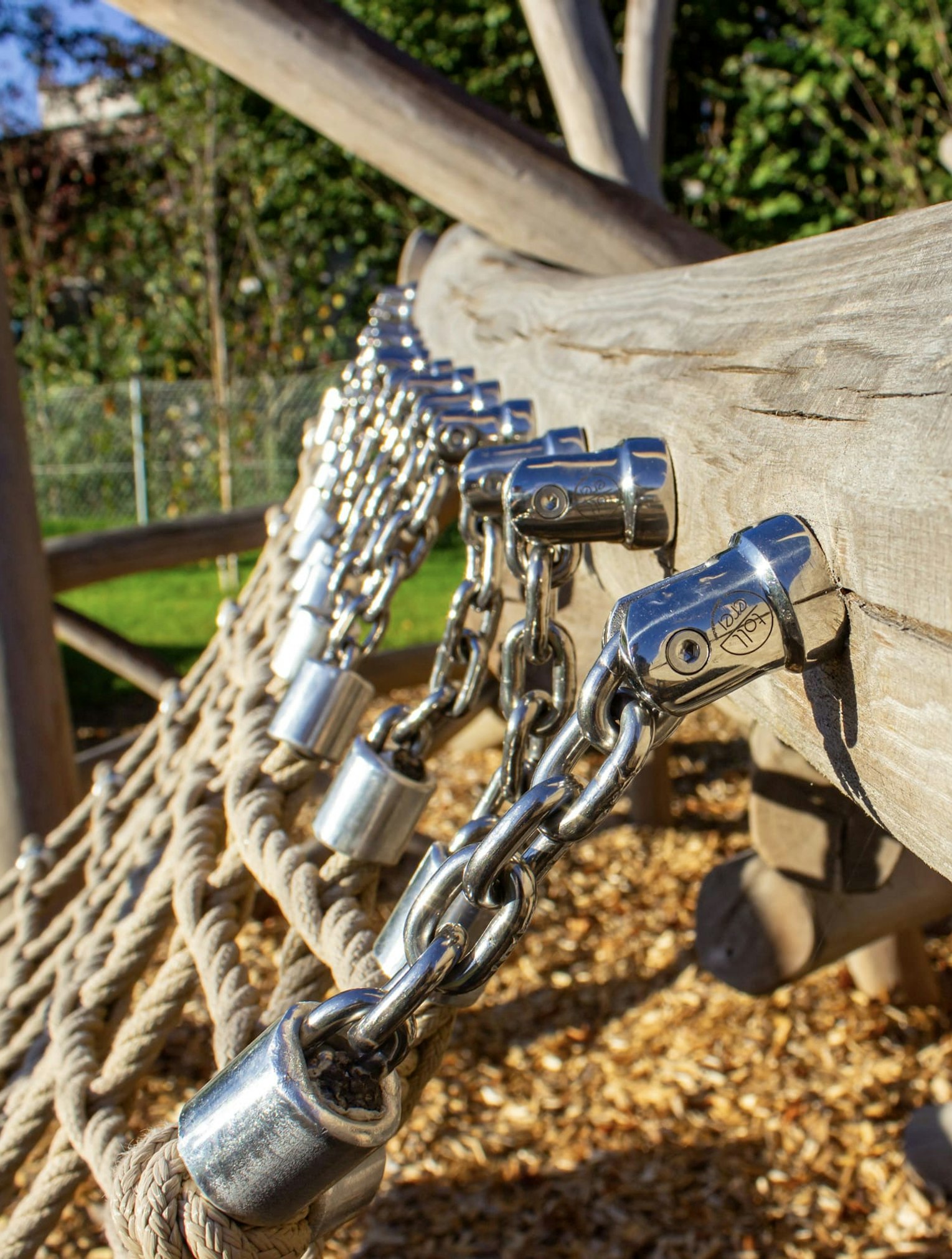 Nahaufnahme von Metallketten, die an einer Holzstruktur befestigt sind, die Teil eines Kletternetzes auf einem Spielplatz ist, mit Holzspänen auf dem Boden.