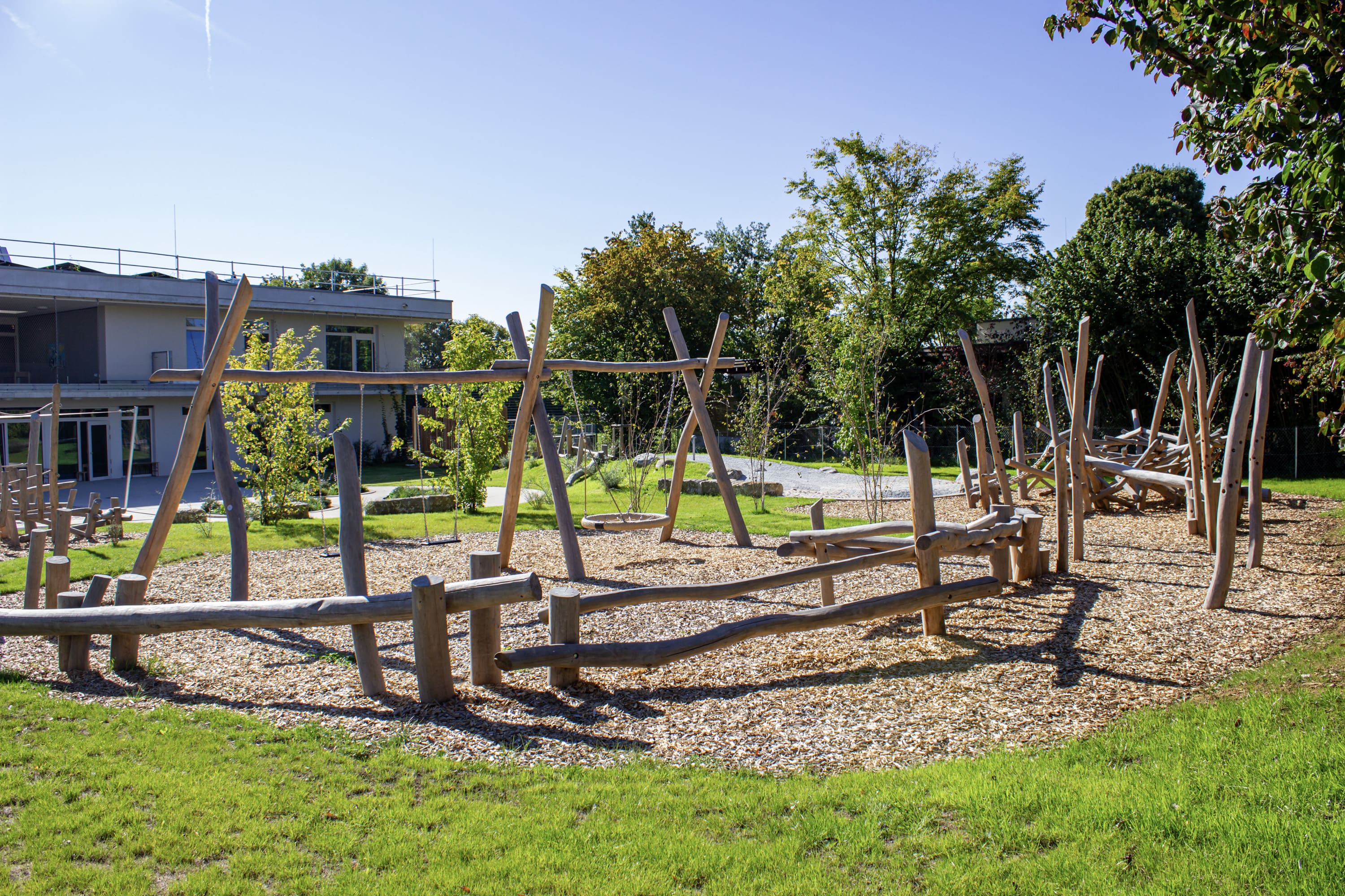 Spielplatz im Freien mit Holzklettergerüsten und Schaukeln, umgeben von Bäumen und einem Gebäude im Hintergrund an einem sonnigen Tag.
