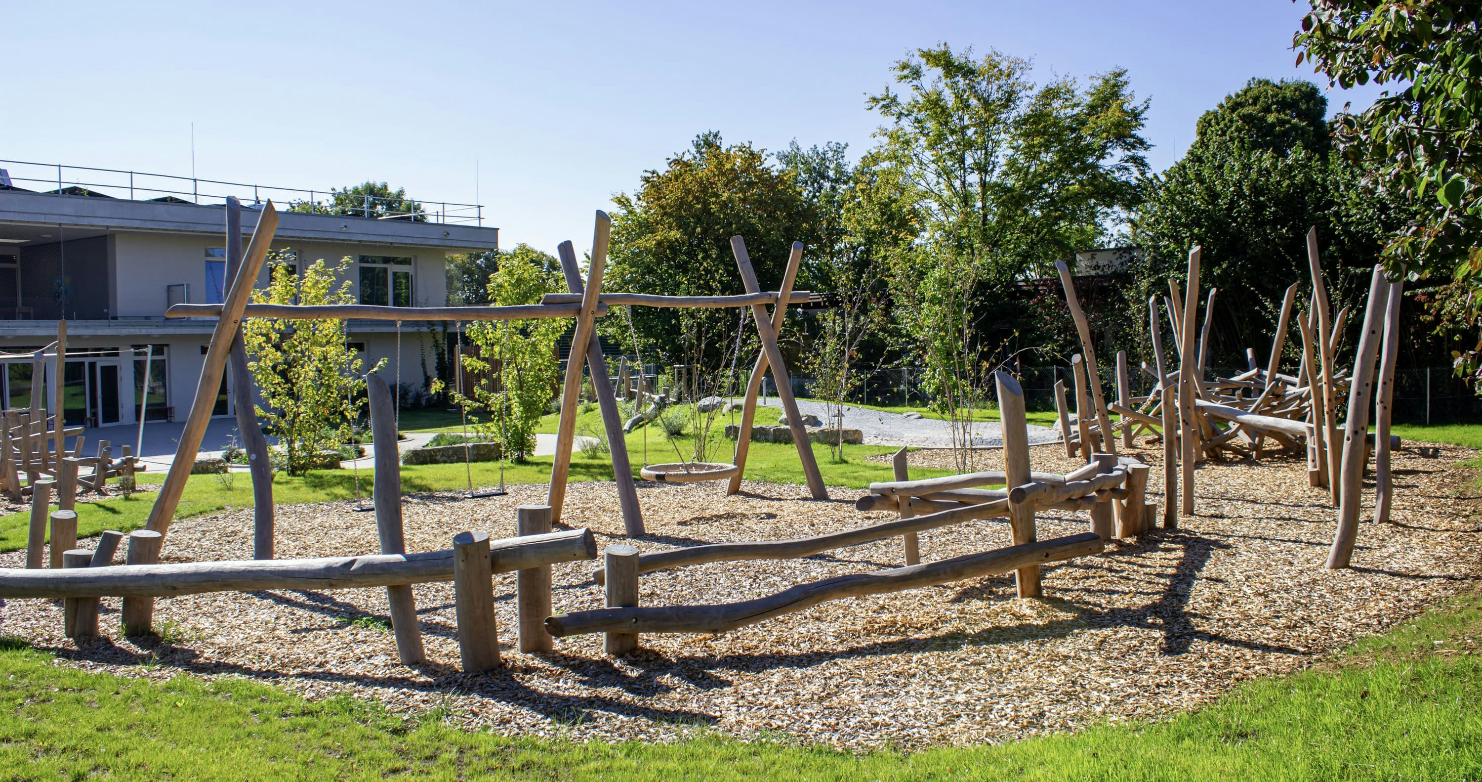 Spielplatz im Freien mit Holzklettergerüsten und Schaukeln, umgeben von Bäumen und einem Gebäude im Hintergrund an einem sonnigen Tag.