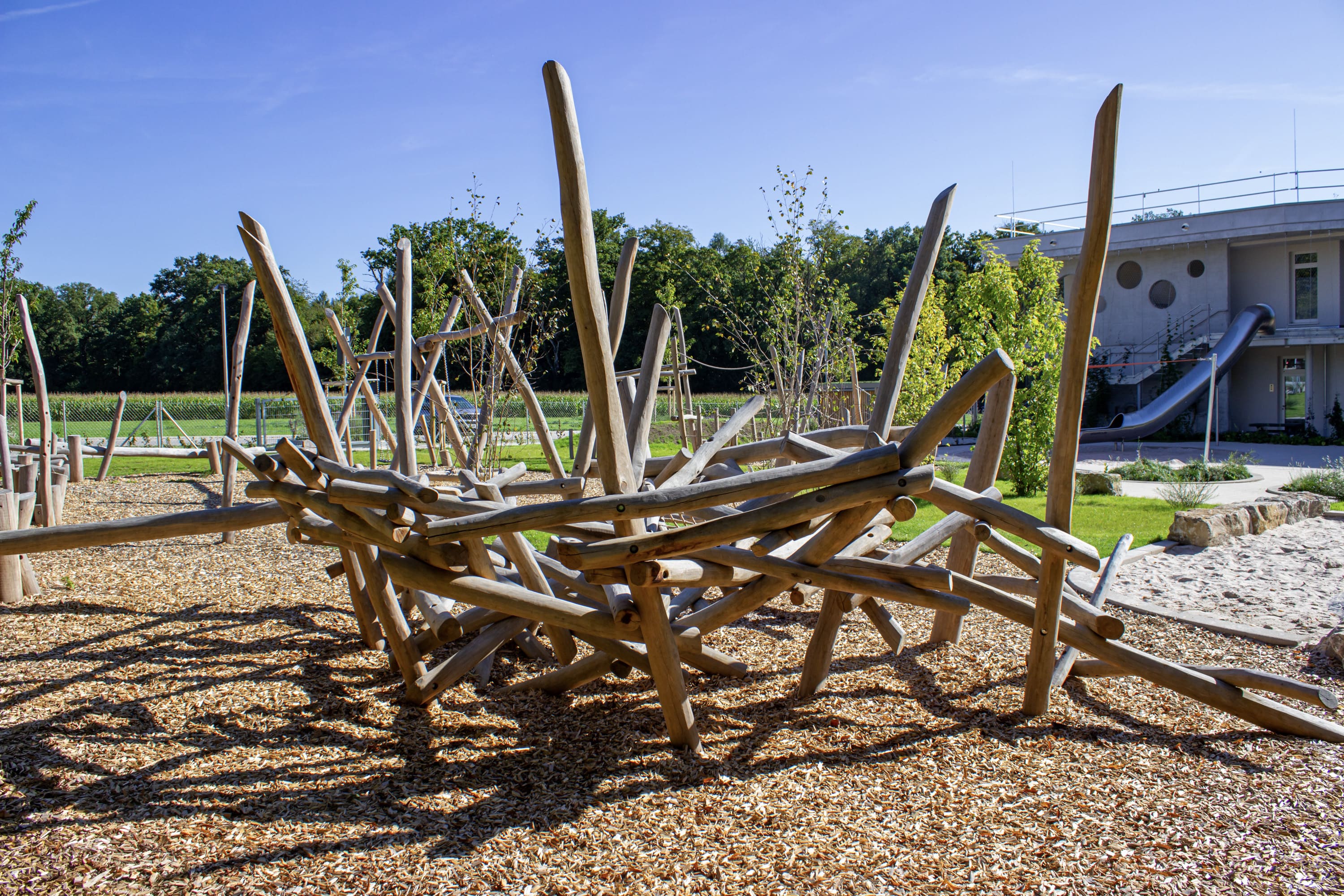 Spielplatz im Freien mit einer abstrakten Kletterstruktur aus Holz auf einer Mulchfläche, umgeben von Gras, Bäumen und einem modernen Gebäude im Hintergrund.