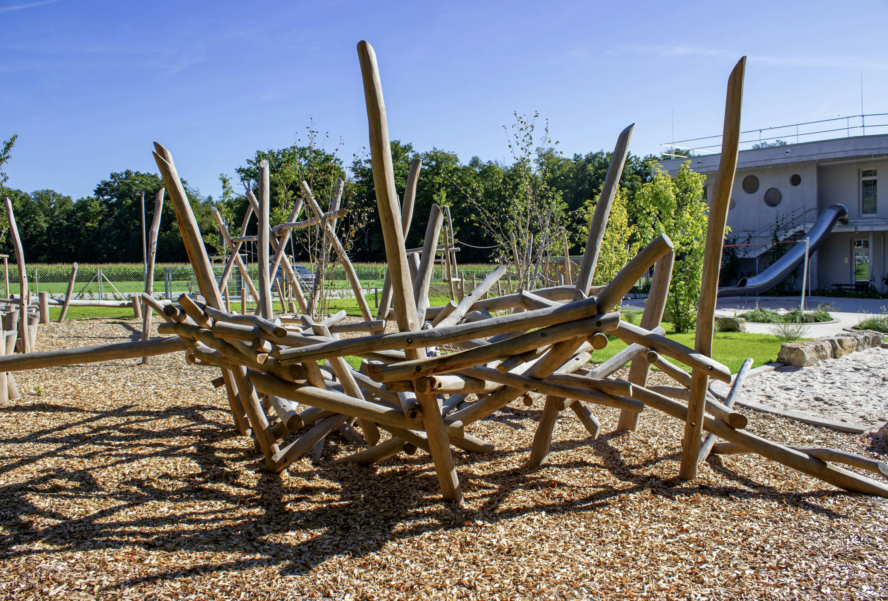 Spielplatz im Freien mit einer abstrakten Kletterstruktur aus Holz auf einer Mulchfläche, umgeben von Gras, Bäumen und einem modernen Gebäude im Hintergrund.