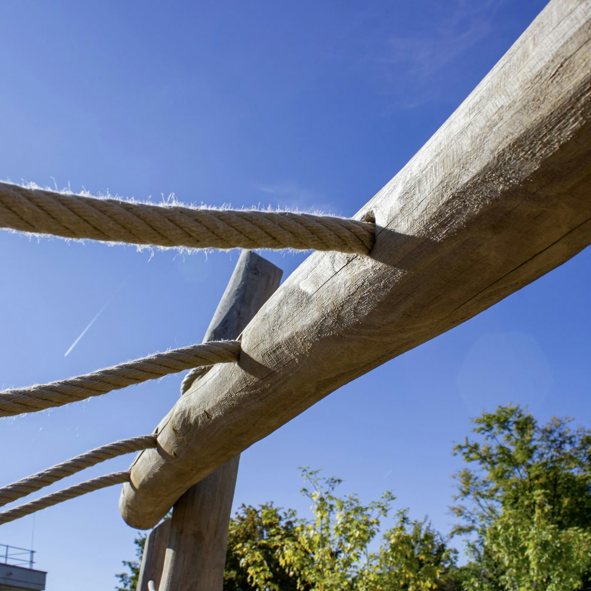 Nahaufnahme von dicken Seilen, die zwischen großen Holzbalken verknotet sind, mit einem klaren blauen Himmel und einigen Bäumen im Hintergrund.