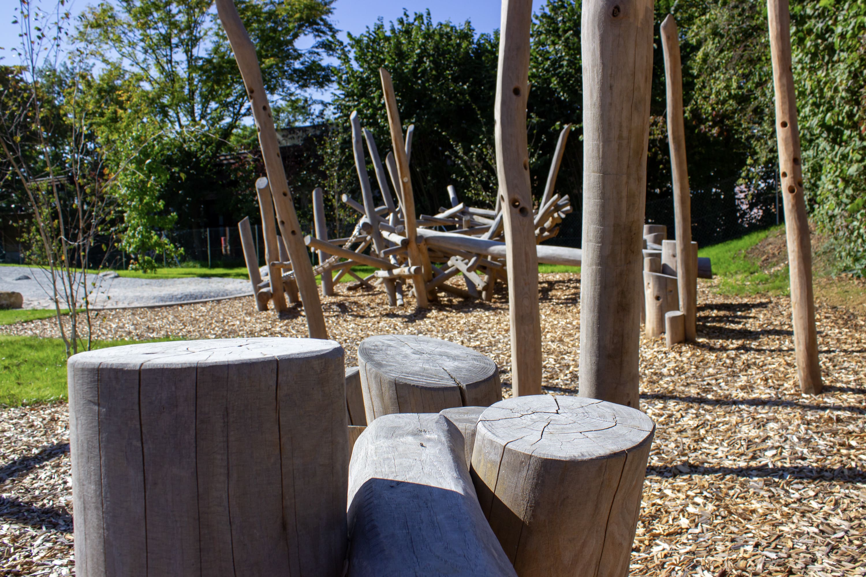 Natürliche Spielgeräte aus Holz und Sitzgelegenheiten aus Holzstämmen auf einem mit Mulch bedeckten Boden, umgeben von Grün und Bäumen in einer Außenanlage.