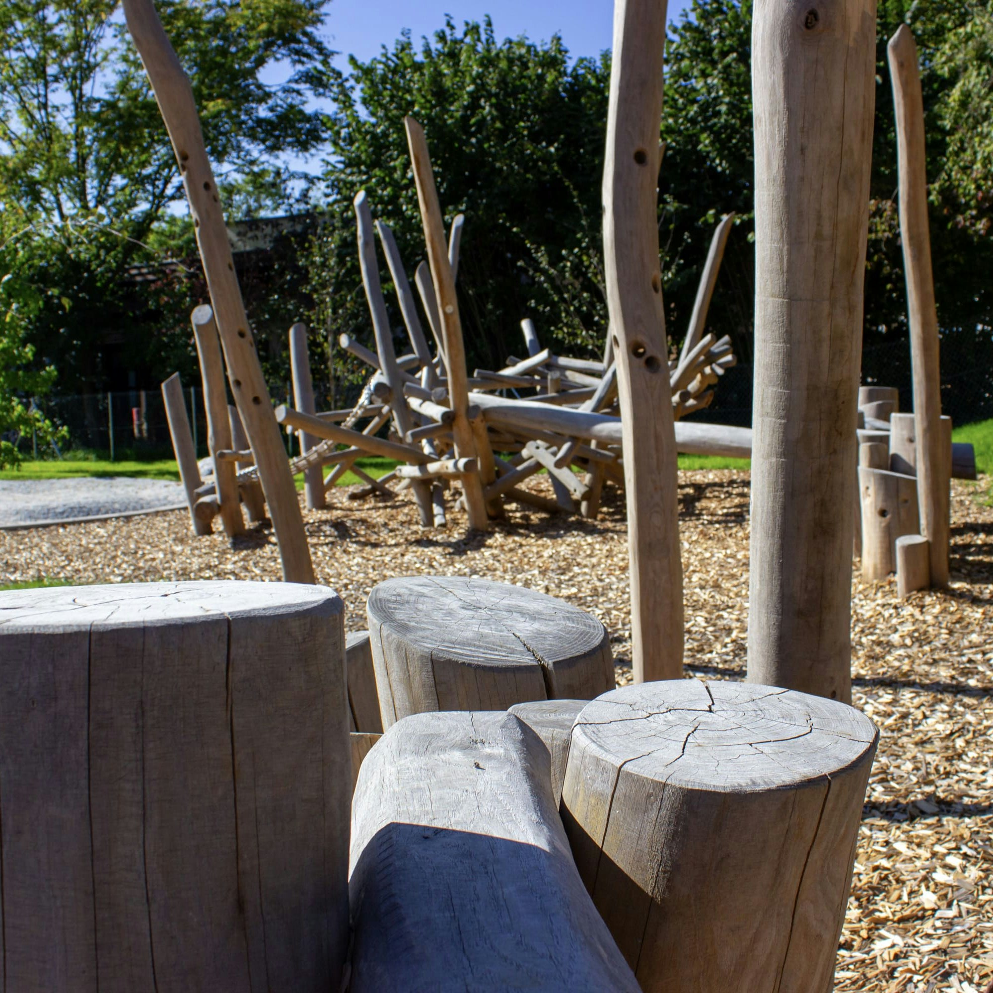 Natürliche Spielgeräte aus Holz und Sitzgelegenheiten aus Holzstämmen auf einem mit Mulch bedeckten Boden, umgeben von Grün und Bäumen in einer Außenanlage.