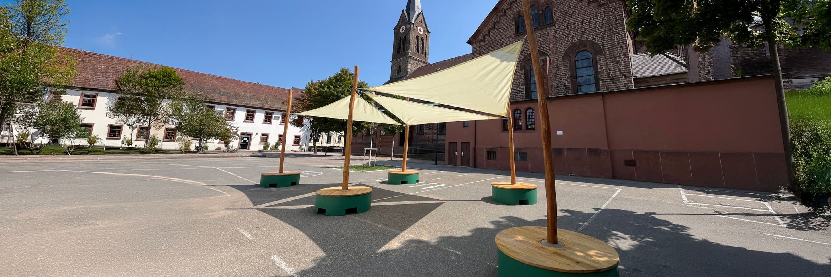 Ein Schulhof mit über Holzpfosten gespannten Sonnensegeln, umgeben von Schulgebäuden und einer Kirche mit einem hohen Uhrenturm im Hintergrund.