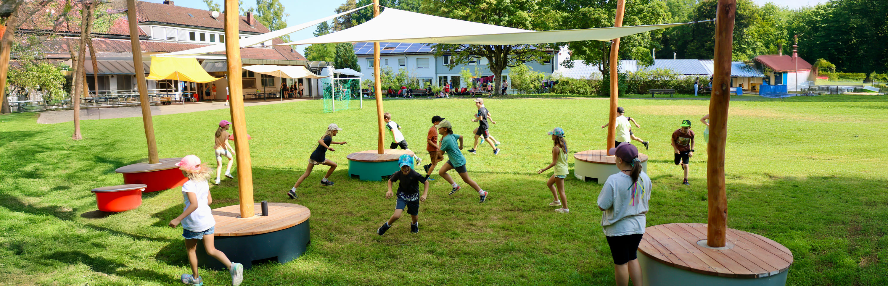 Kinder rennen und spielen im Freien unter Schattensegeln auf einem grasbewachsenen Schulhof, mit Gebäuden und Bäumen im Hintergrund.