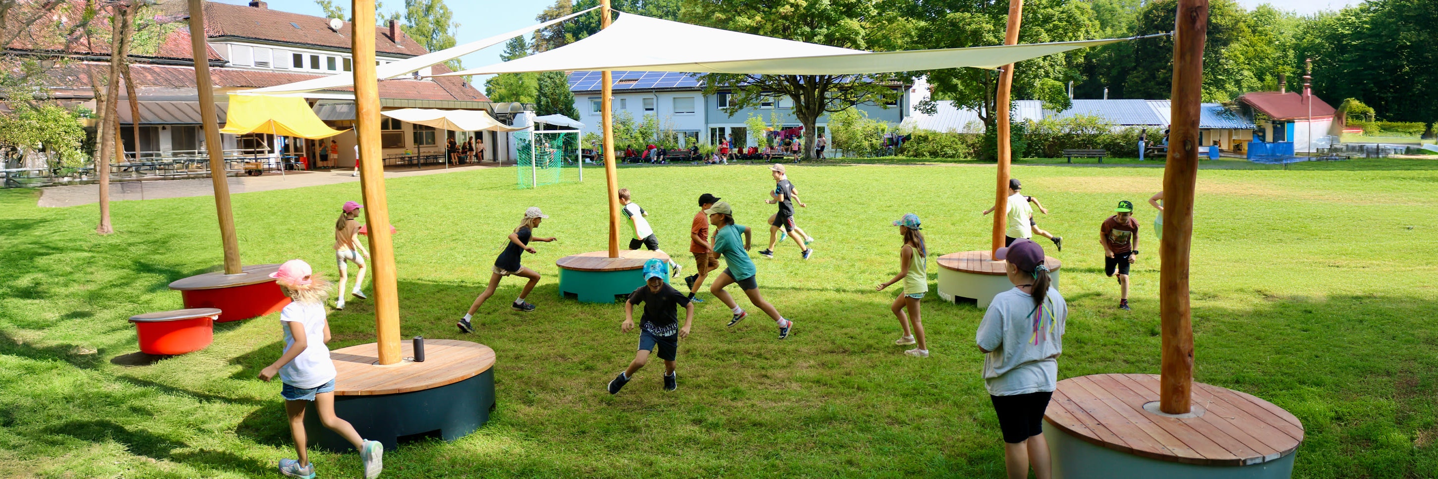 Kinder rennen und spielen im Freien unter Schattensegeln auf einem grasbewachsenen Schulhof, mit Gebäuden und Bäumen im Hintergrund.