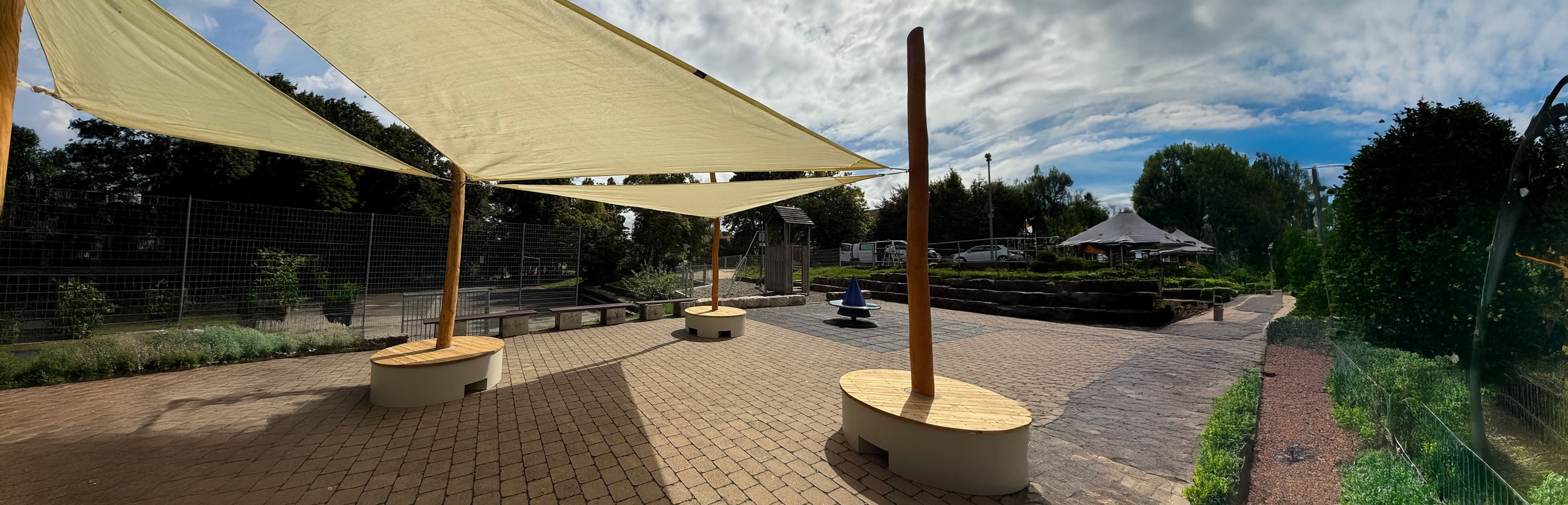 Terrassenbereich mit großen Sonnensegeln, runden Holzbänken, Ziegelsteinpflaster, Grünpflanzen und einem teilweise bewölkten Himmel.